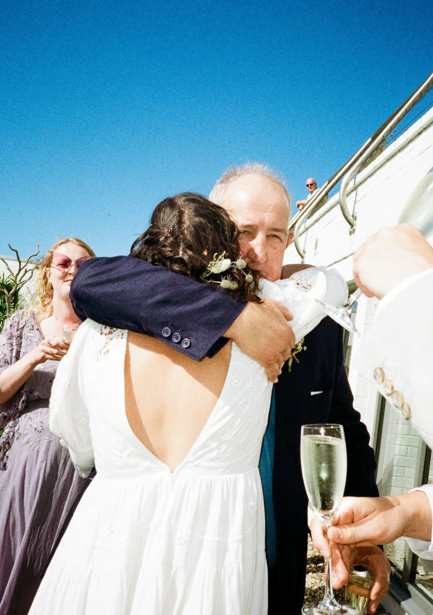 Wedding celebration outdoors with guests in formal attire against white building backdrop.