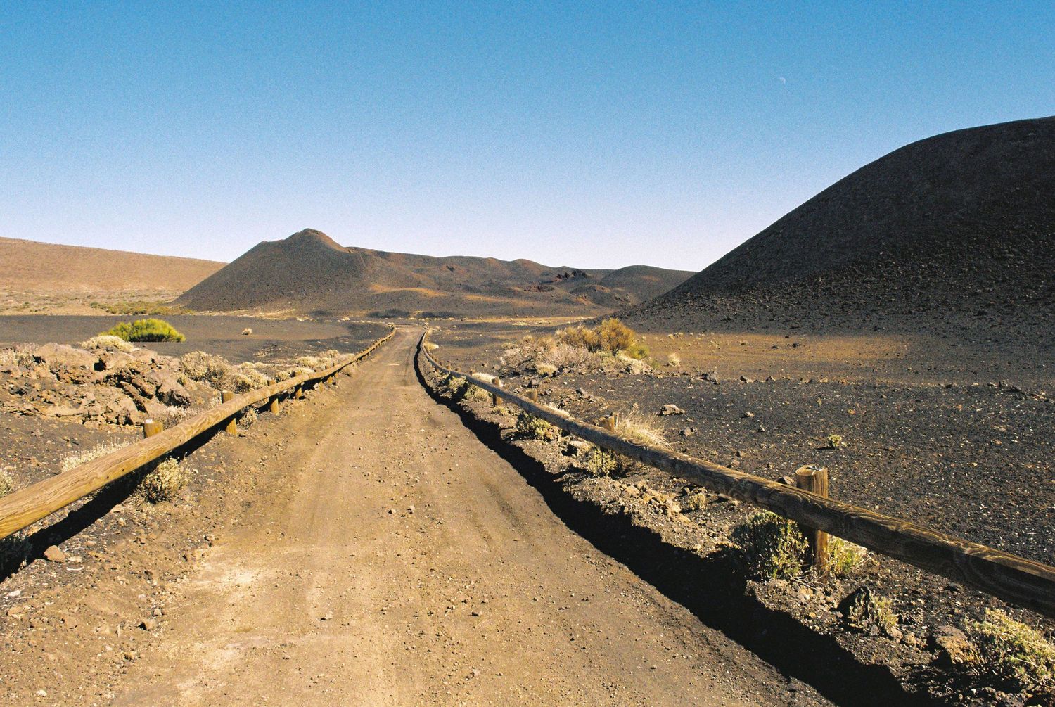 Winding dirt road cuts through a vast desert landscape under clear blue skies.