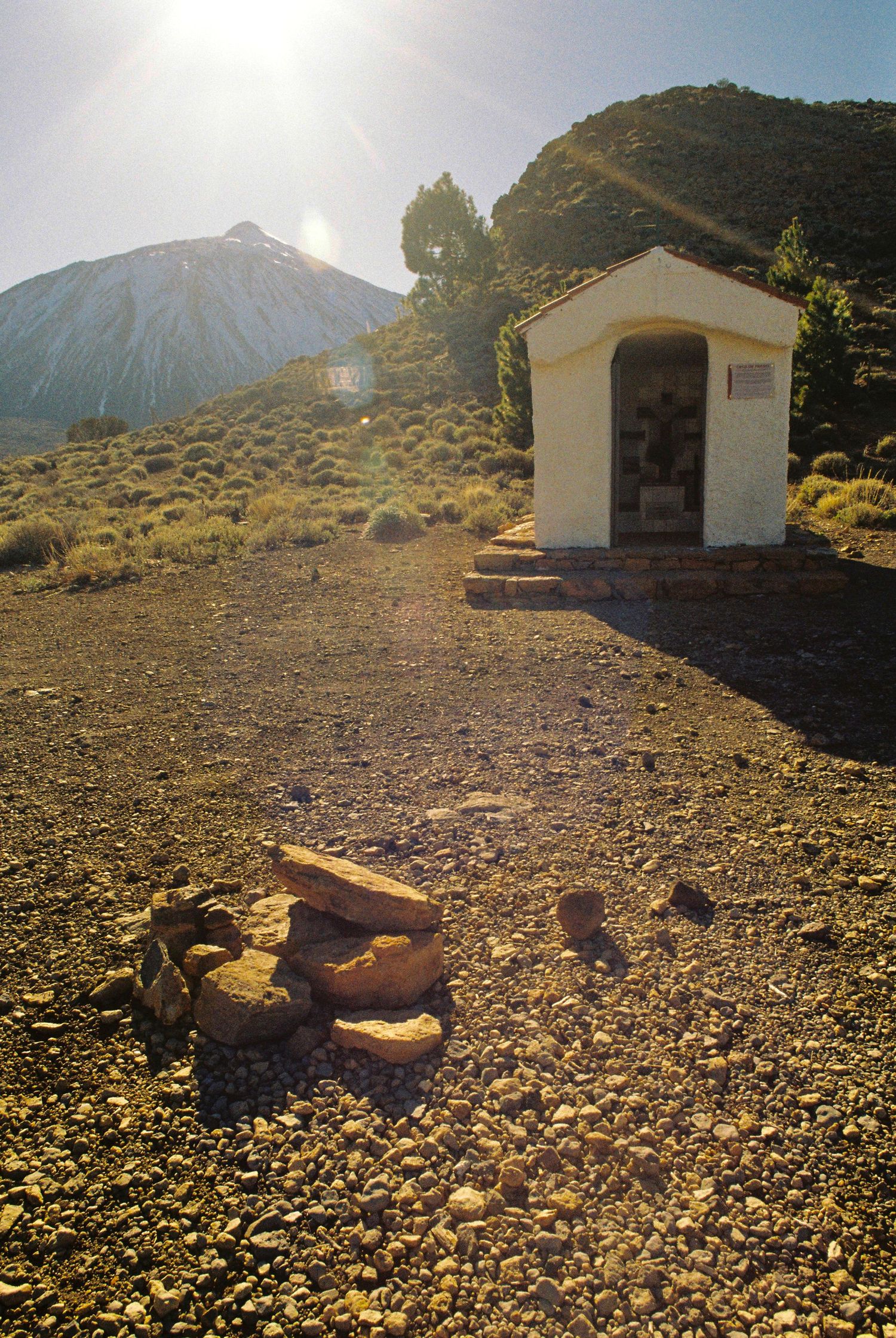 Small white building sits on rocky terrain with mountains in the background under bright sunlight.