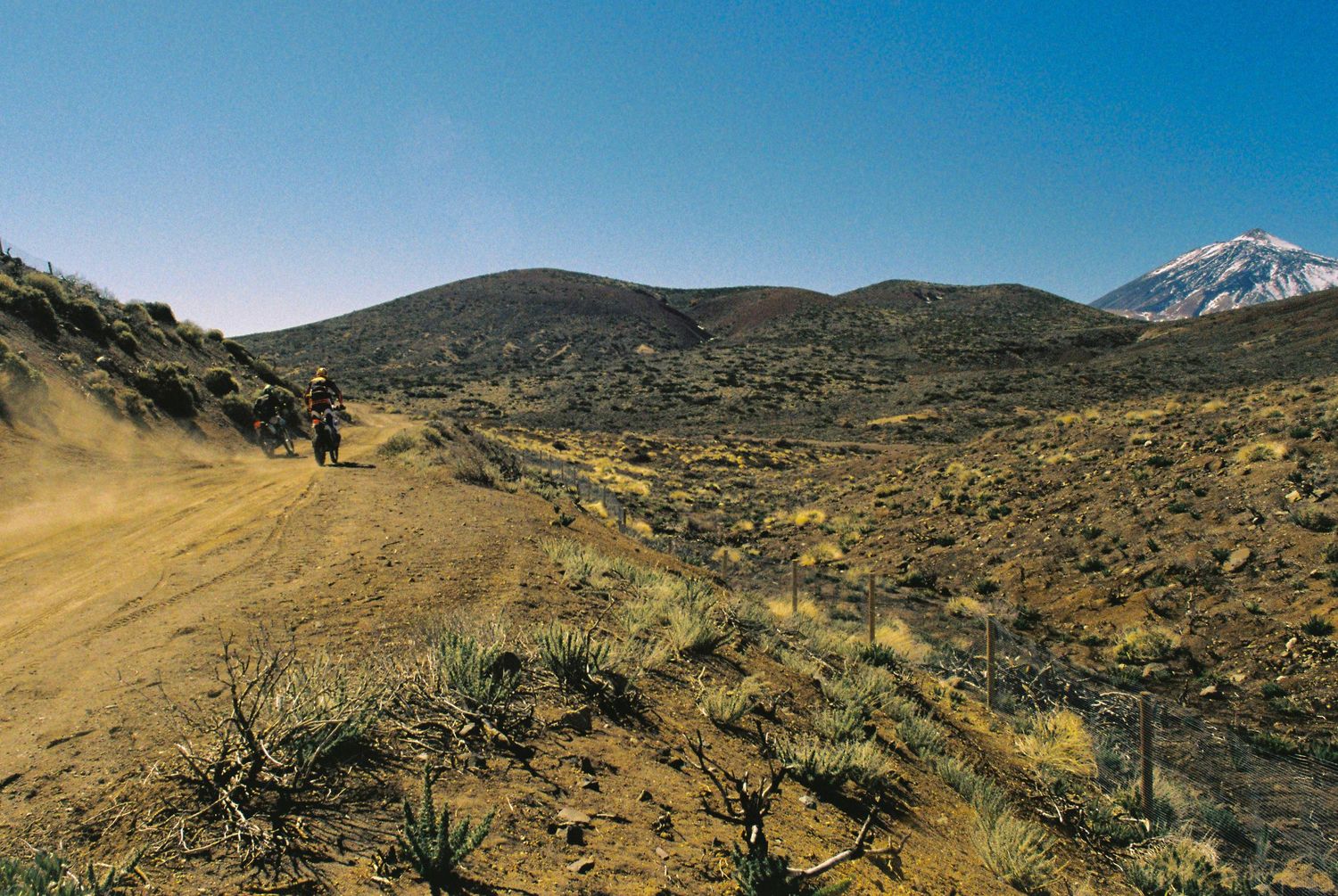 A panoramic view of a rugged mountain trail winding through arid terrain under bright blue skies.
