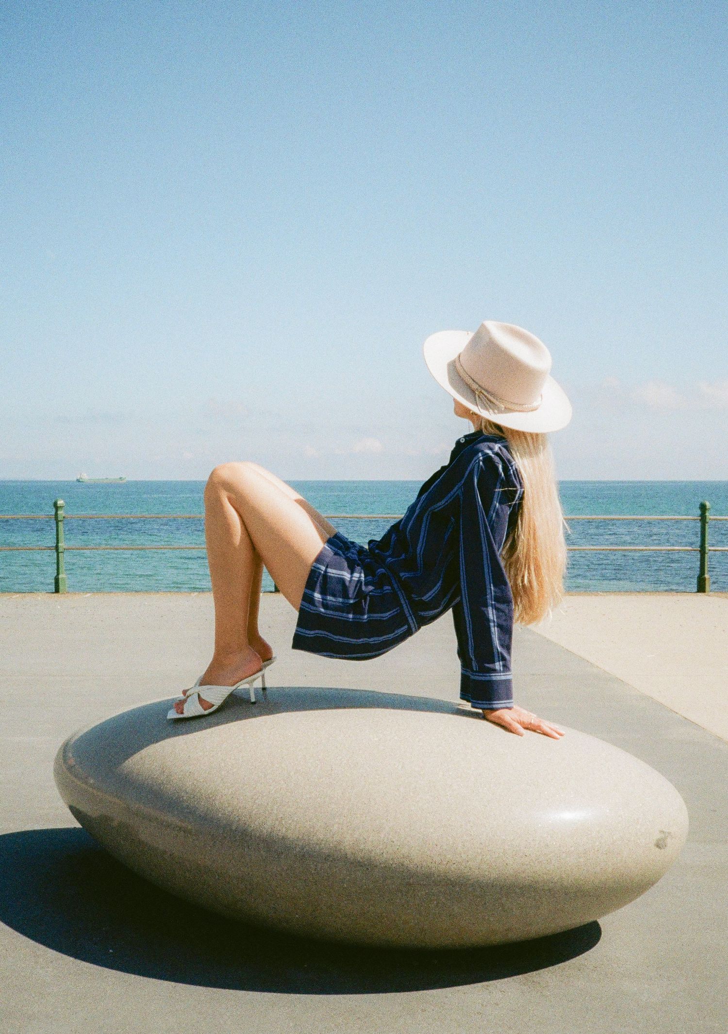 A figure in a striped shirt and sun hat reclines on a curved concrete structure by the ocean.