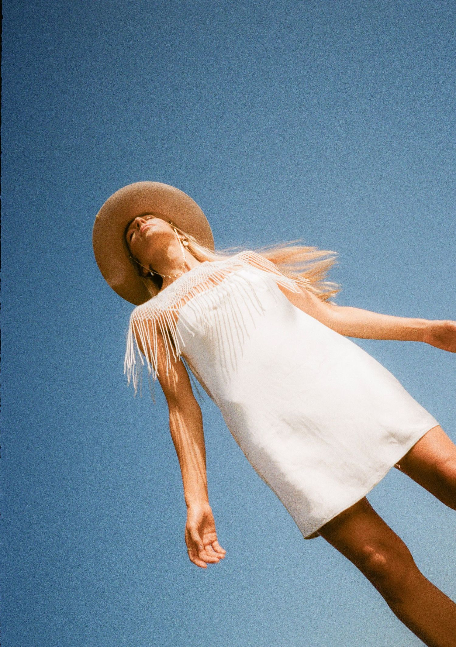 Artistic fashion series with flowing white dress and brown hat against blue sky background.