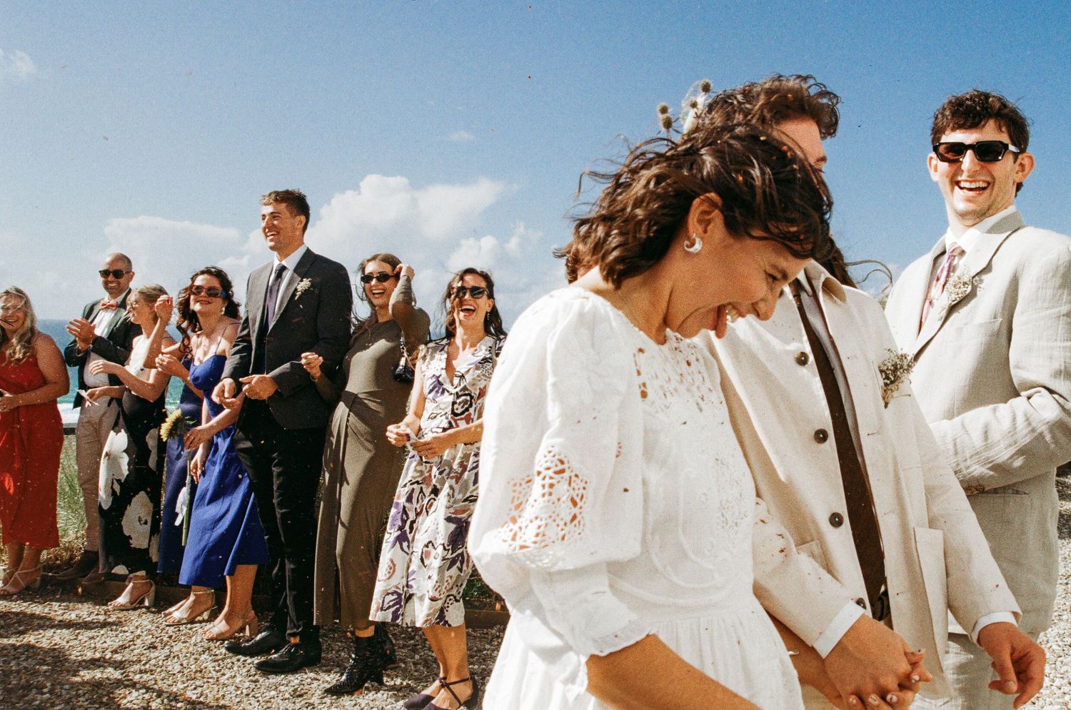Wedding guests stand in rows outdoors under bright blue sky during the ceremony.