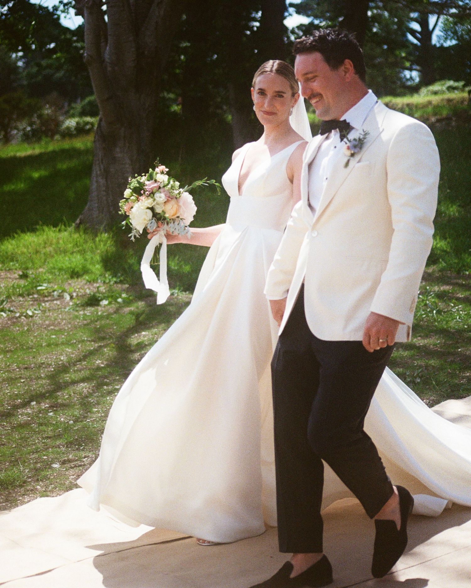 Wedding couple walks together on grass with flowing white dress trailing behind.