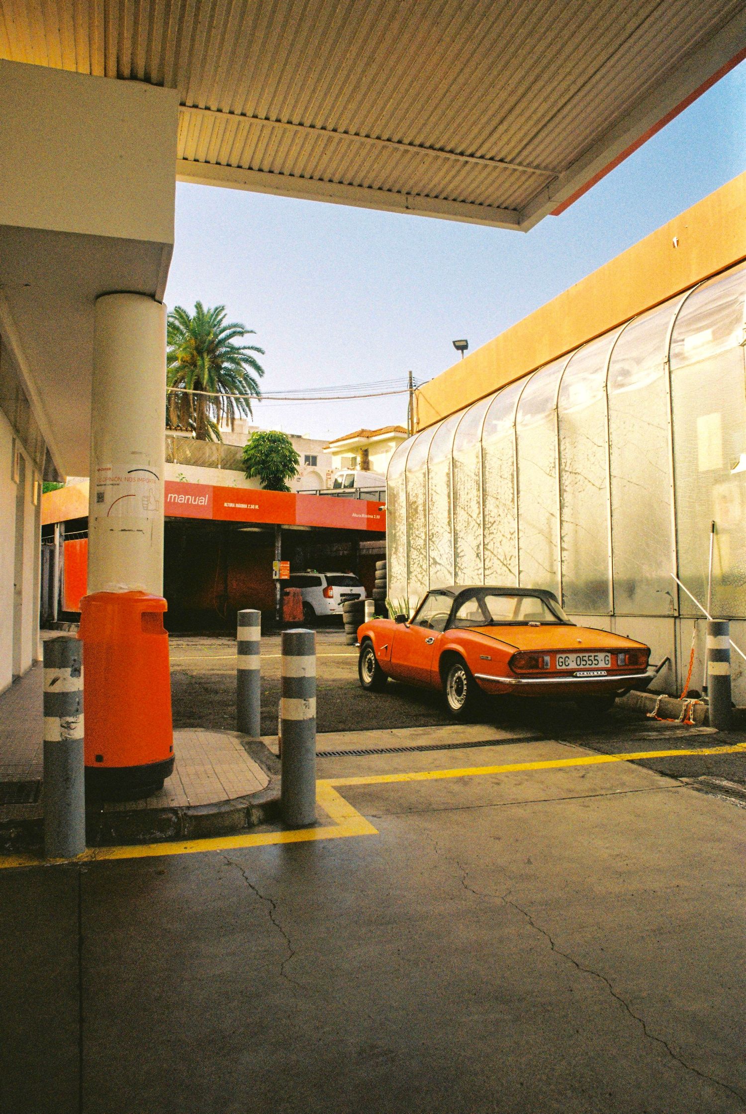 Orange vintage car parked under covered parking area with white walls and concrete pillars.