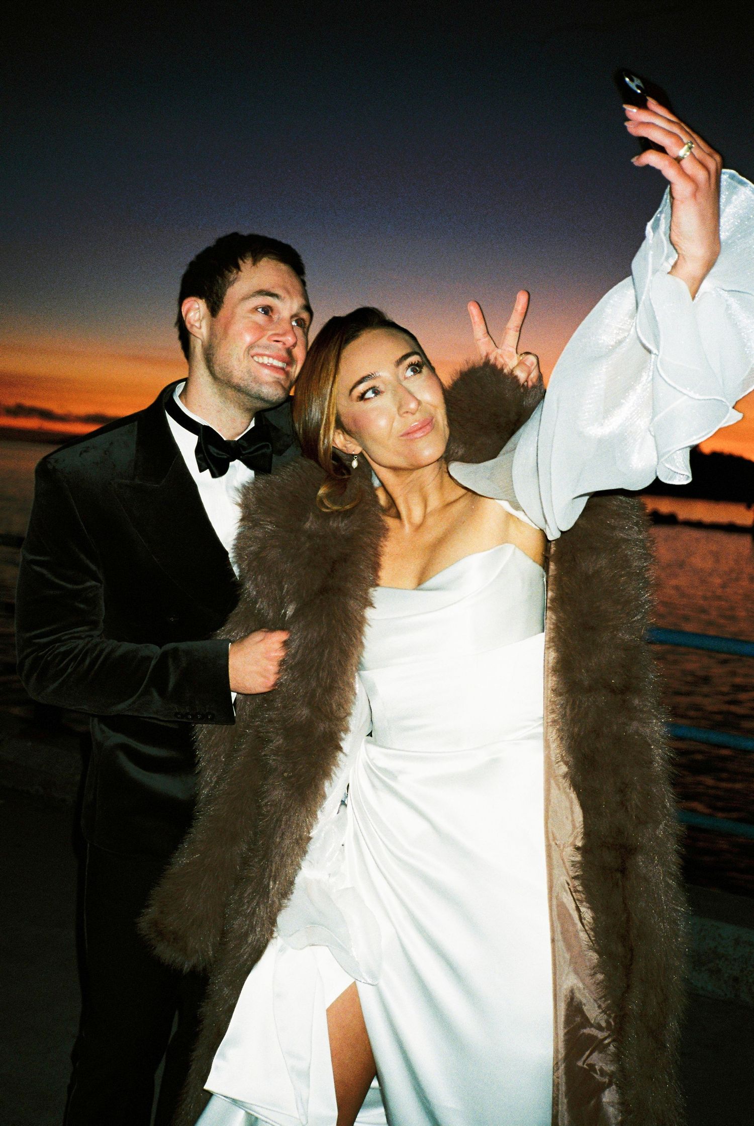 Couple in formal attire poses playfully with fur stoles against a sunset backdrop.
