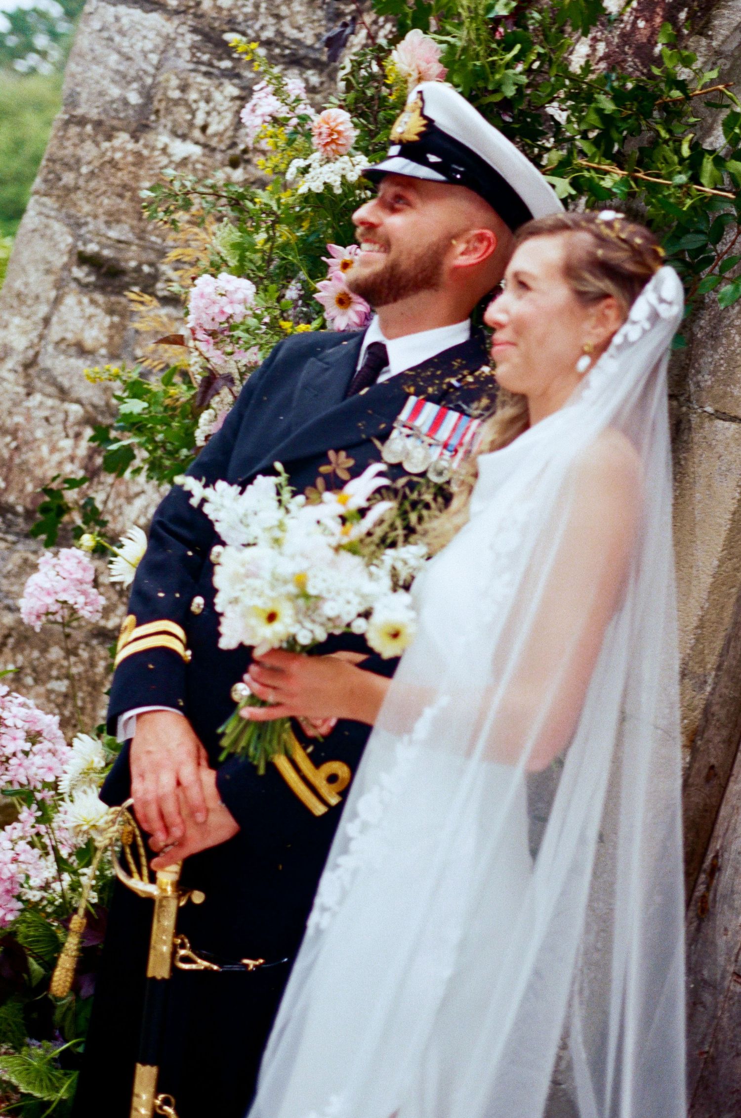 Military officer and bride share joyful laughter during outdoor wedding ceremony against stone wall backdrop.