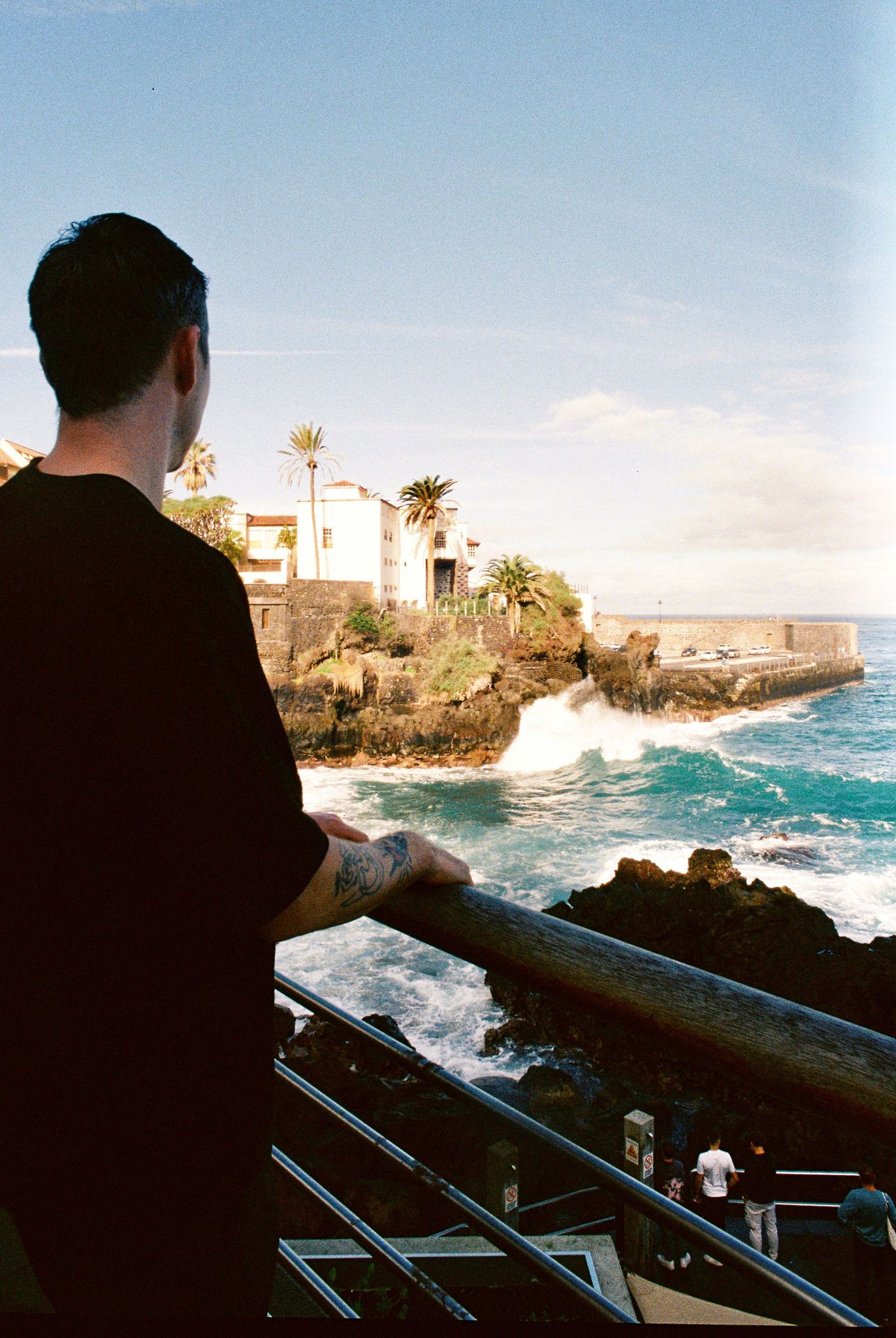 Dramatic coastal seascape with crashing waves against rocky shoreline viewed through metal railings at sunset.