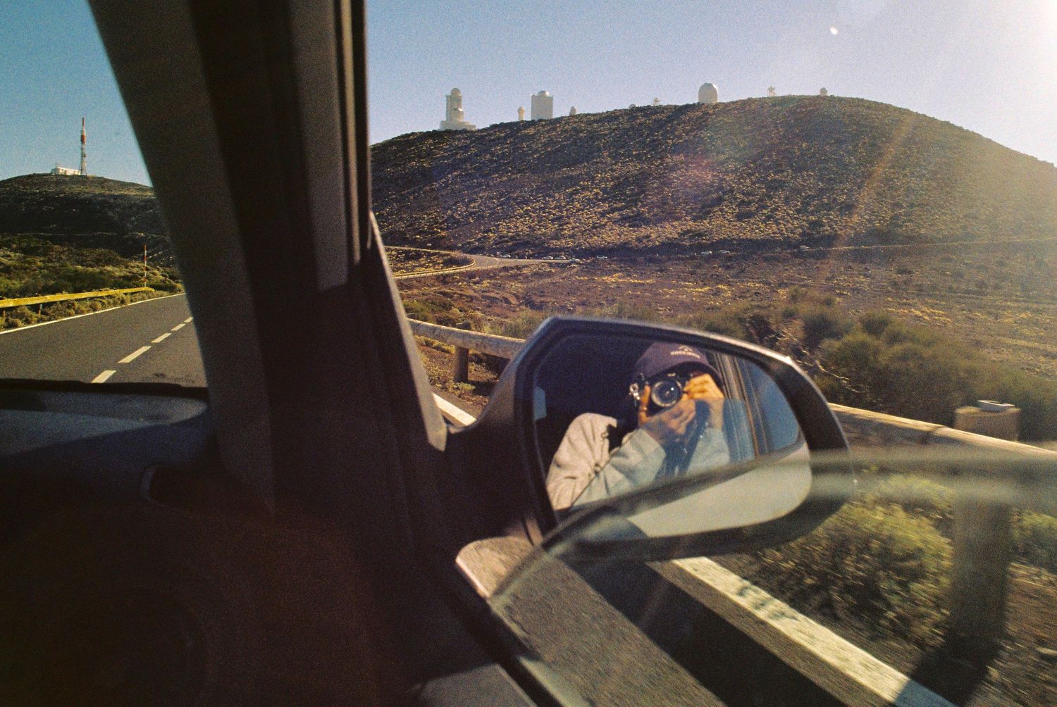 Desert highway stretches into distance with mountains and blue sky visible through car window.