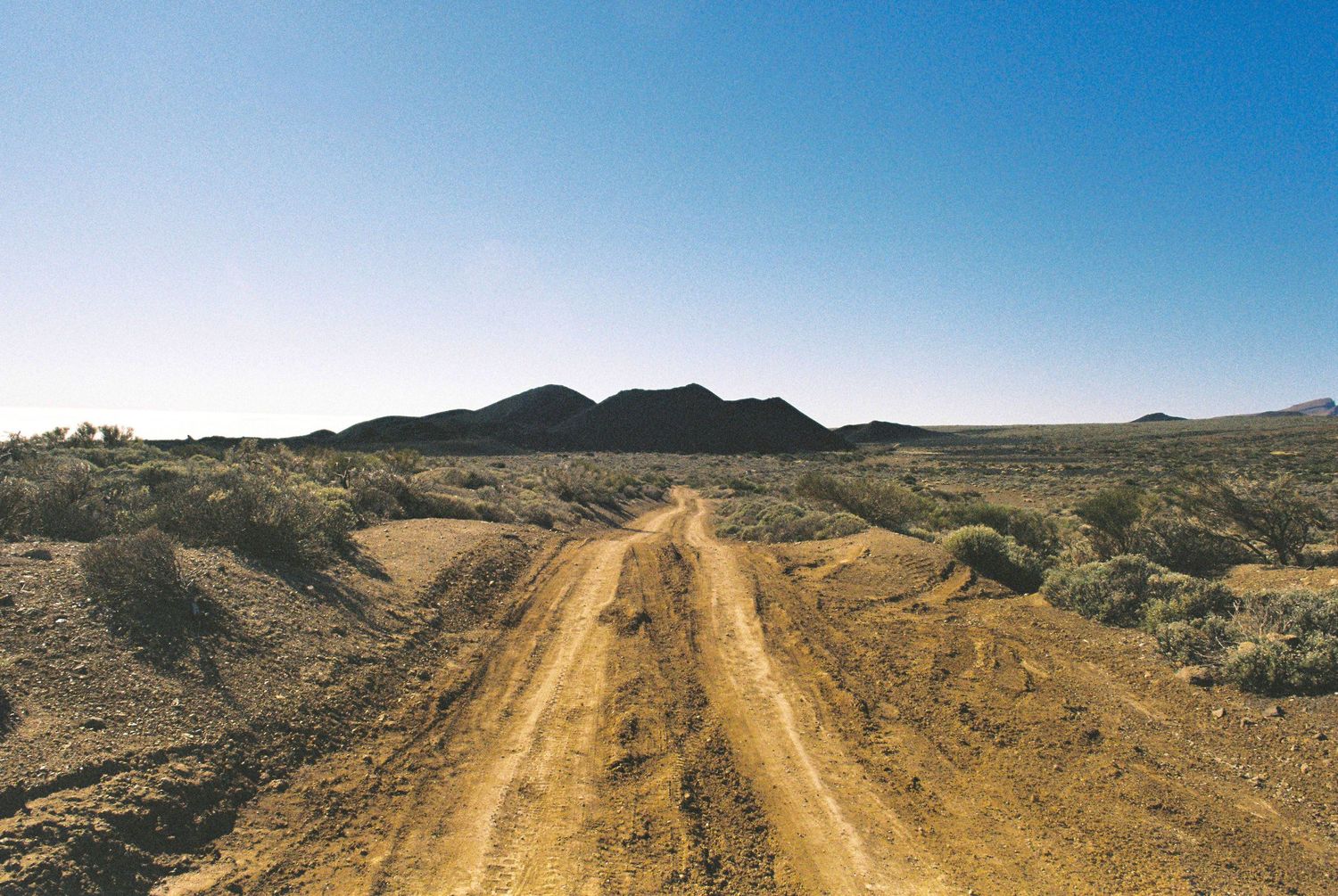 Dusty trail winds through arid desert terrain with mountains in the distance.