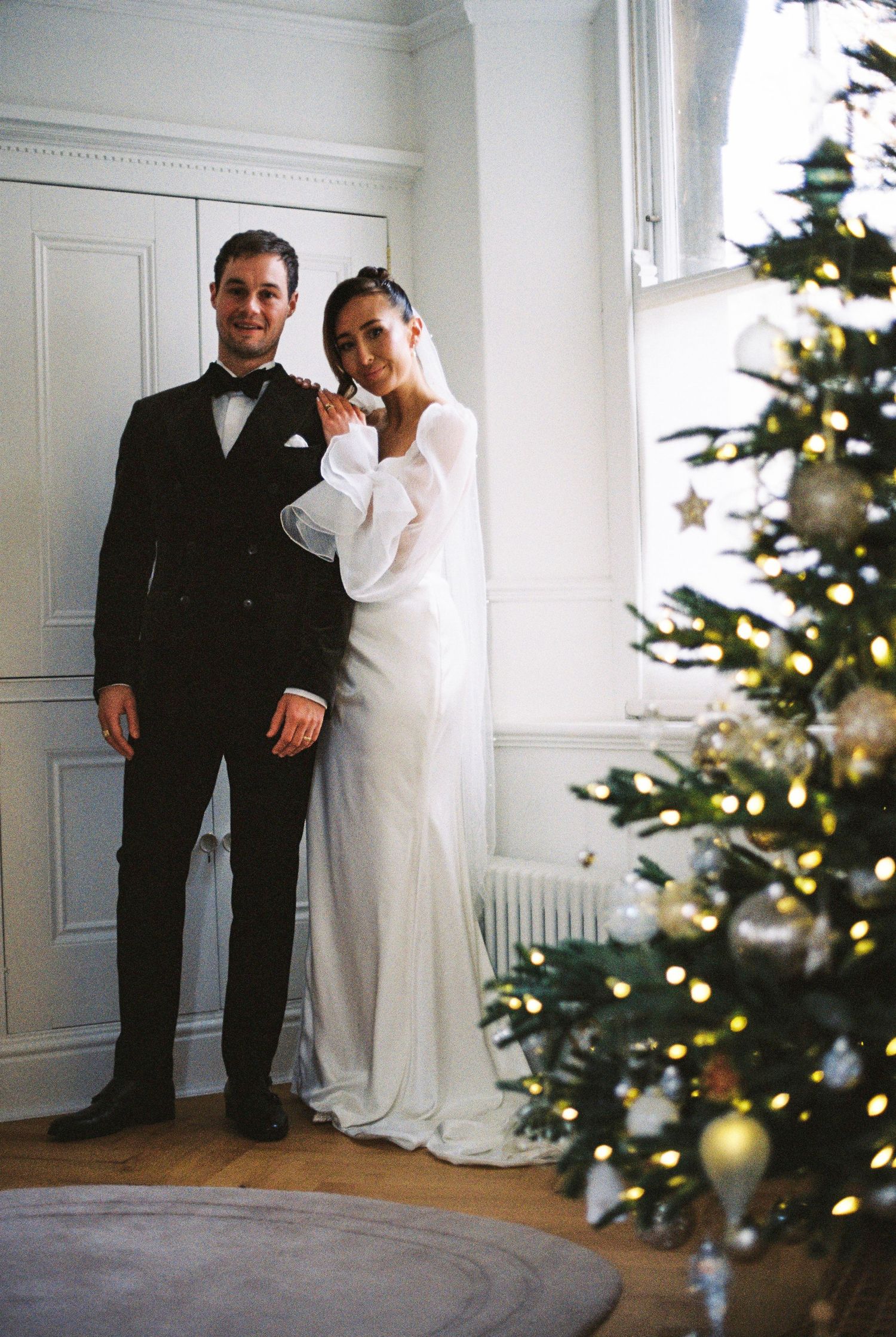 A couple poses together near a decorated Christmas tree in a bright white room during their winter wedding celebration.