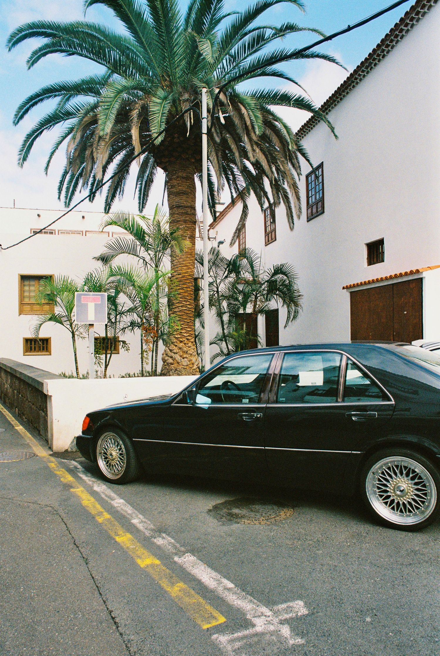 Black vintage car parked on a street with palm trees and white buildings in Mediterranean style architecture.