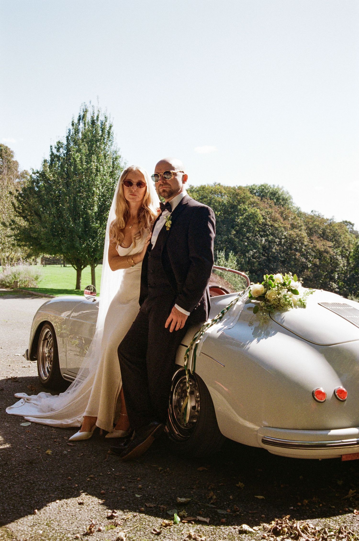 Newlyweds pose beside a classic silver vintage car on their wedding day.