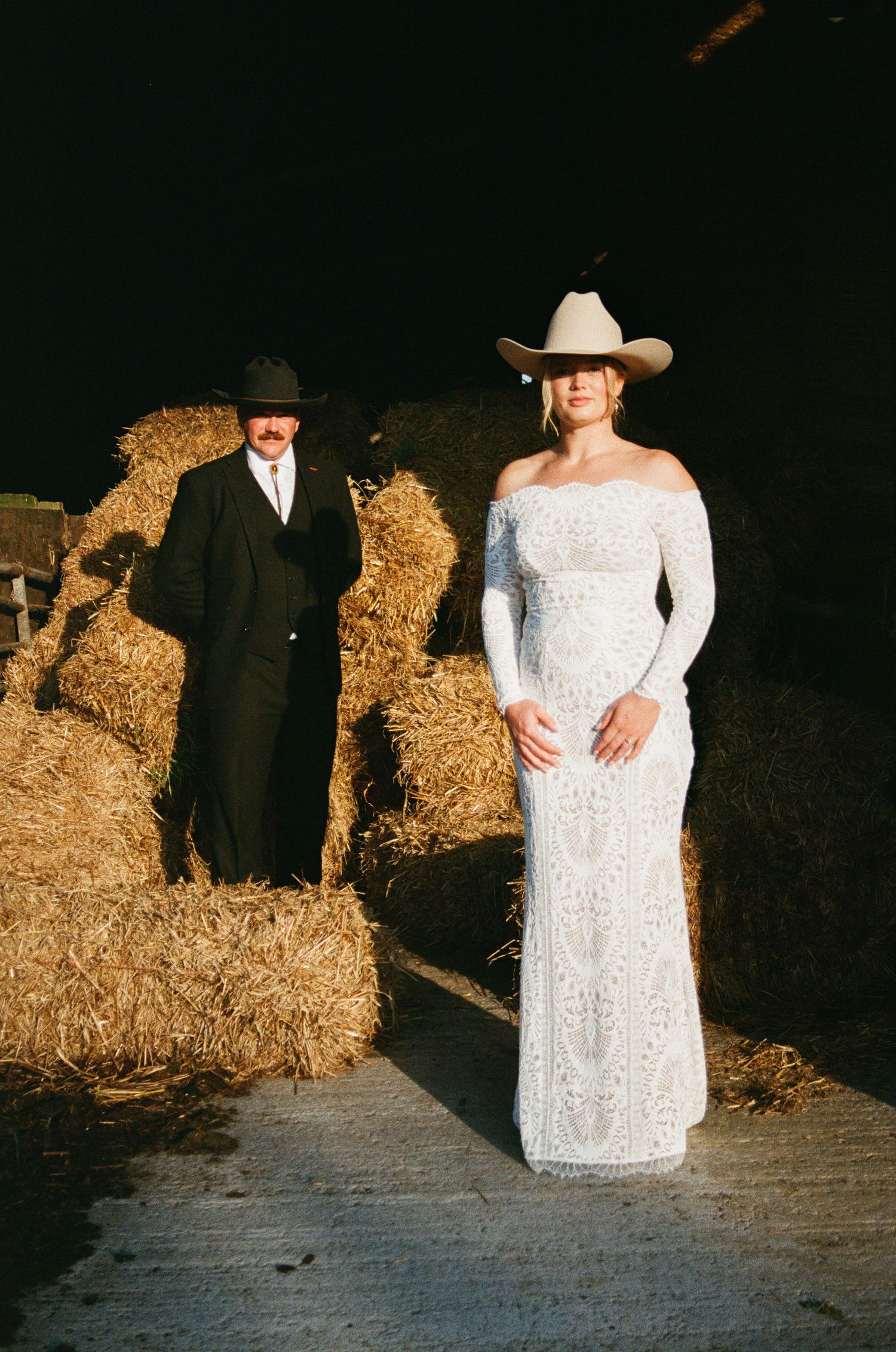 A couple poses near hay bales in a rustic barn setting with dramatic lighting and western attire.