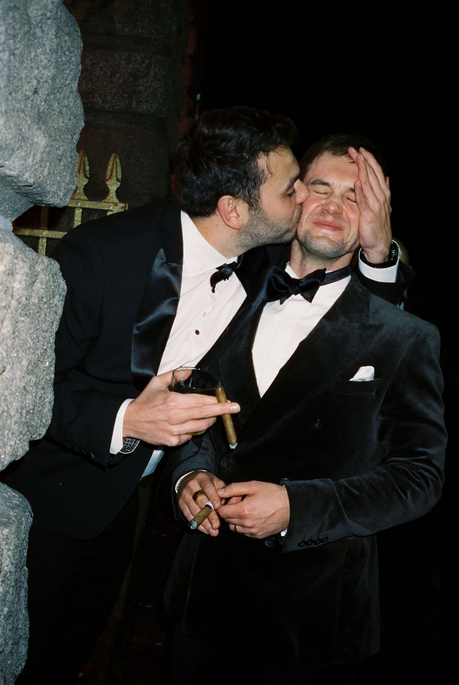 Two people in formal black tuxedos and bow ties sharing cigars in dark setting.