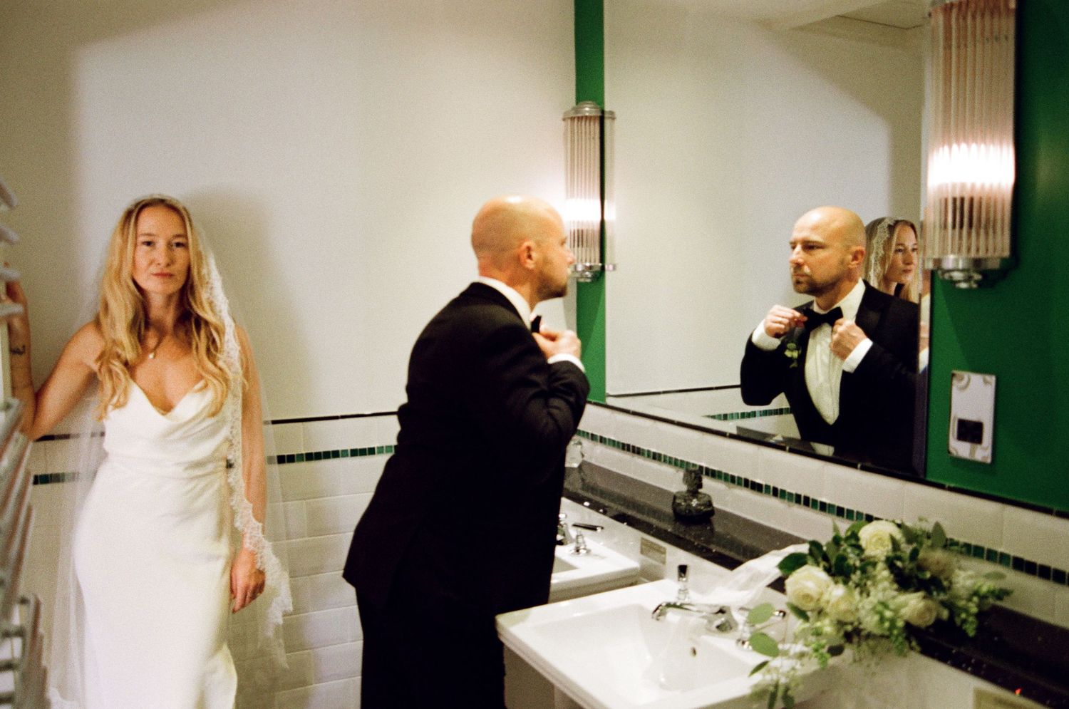 A wedding couple shares a moment in front of bathroom mirrors with floral decorations.
