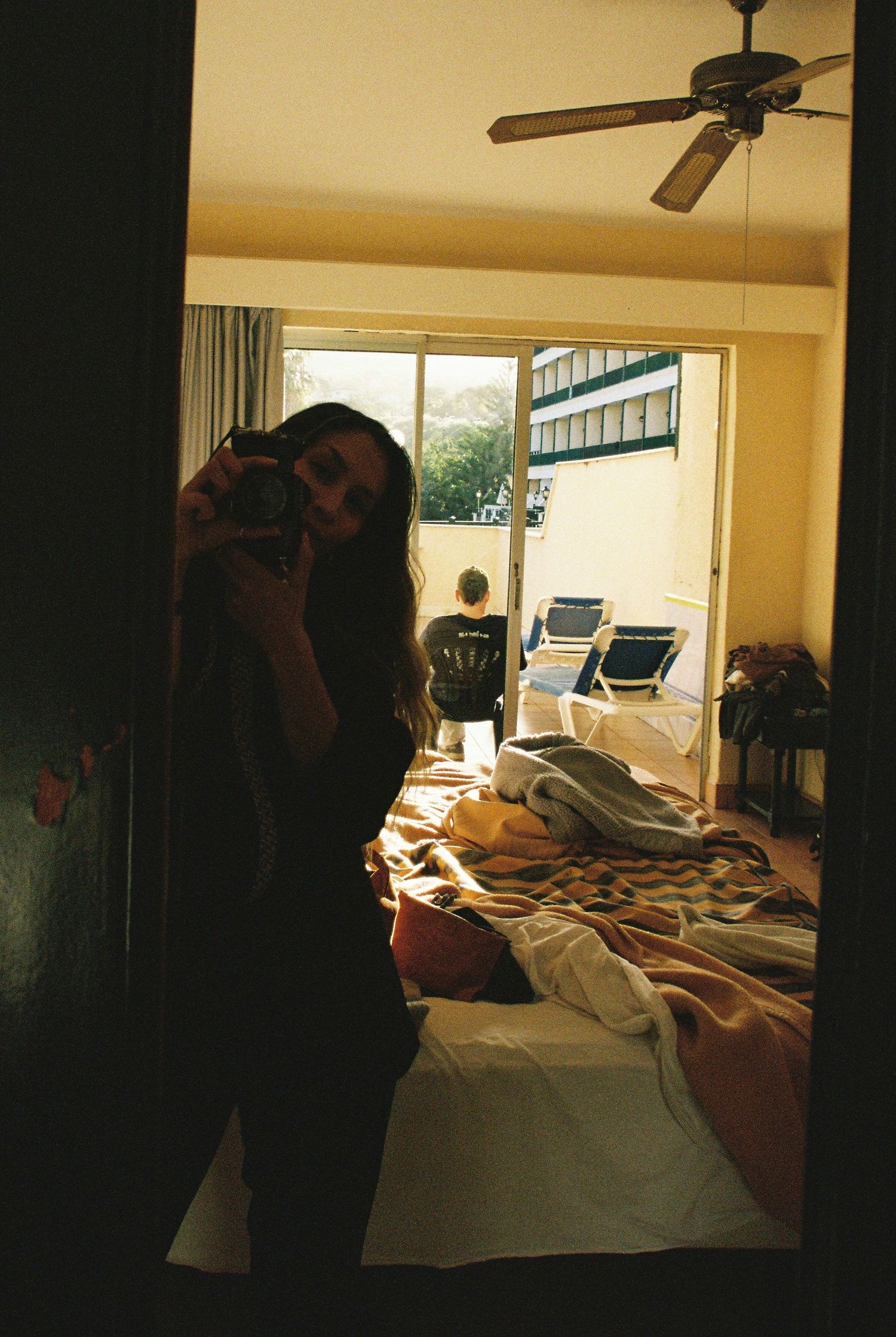 A dimly lit bedroom interior with unmade bed and ceiling fan, showing someone taking a photo in the foreground.