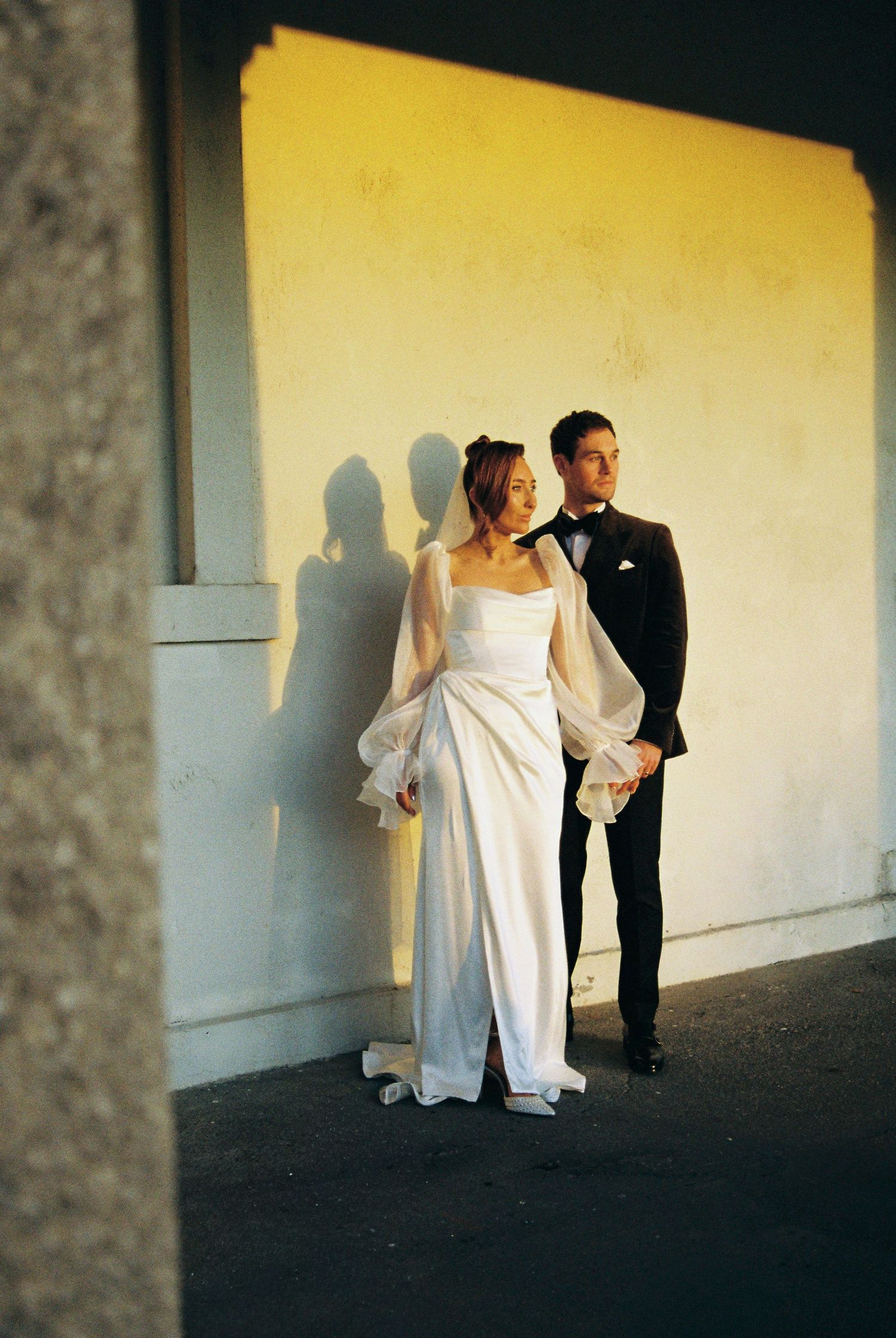 A wedding couple poses against a light wall creating dramatic shadows in an artistic composition.