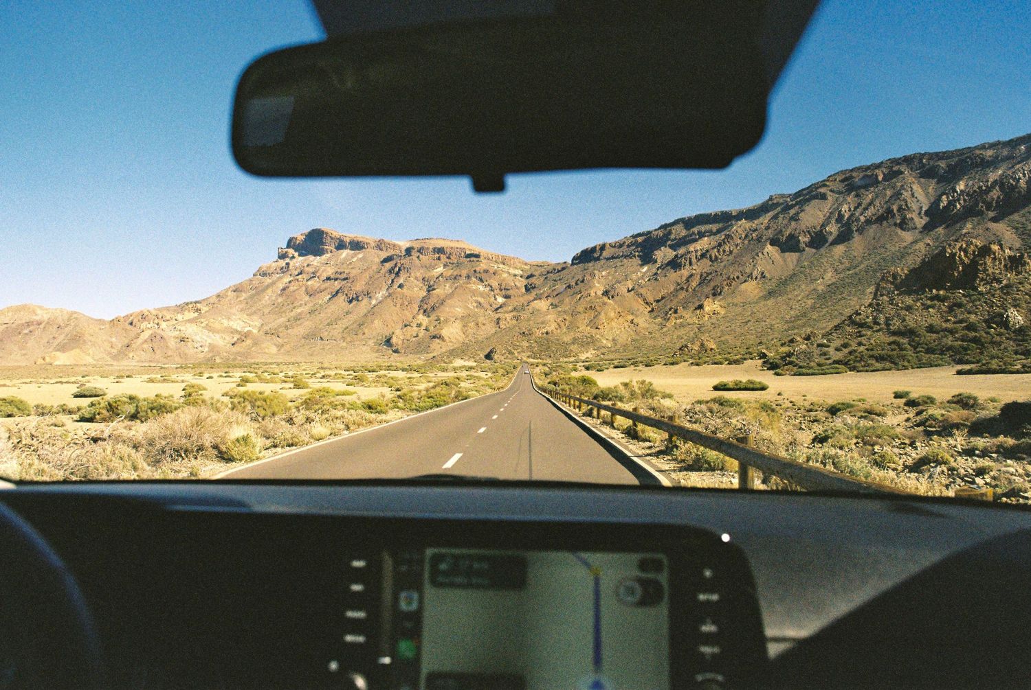 Desert highway stretches into the distance with mountains visible through car windshield.