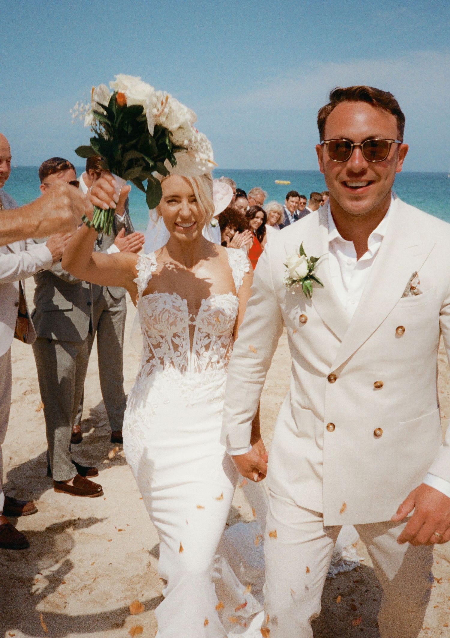 Wedding party in white attire celebrating on sunny beach.