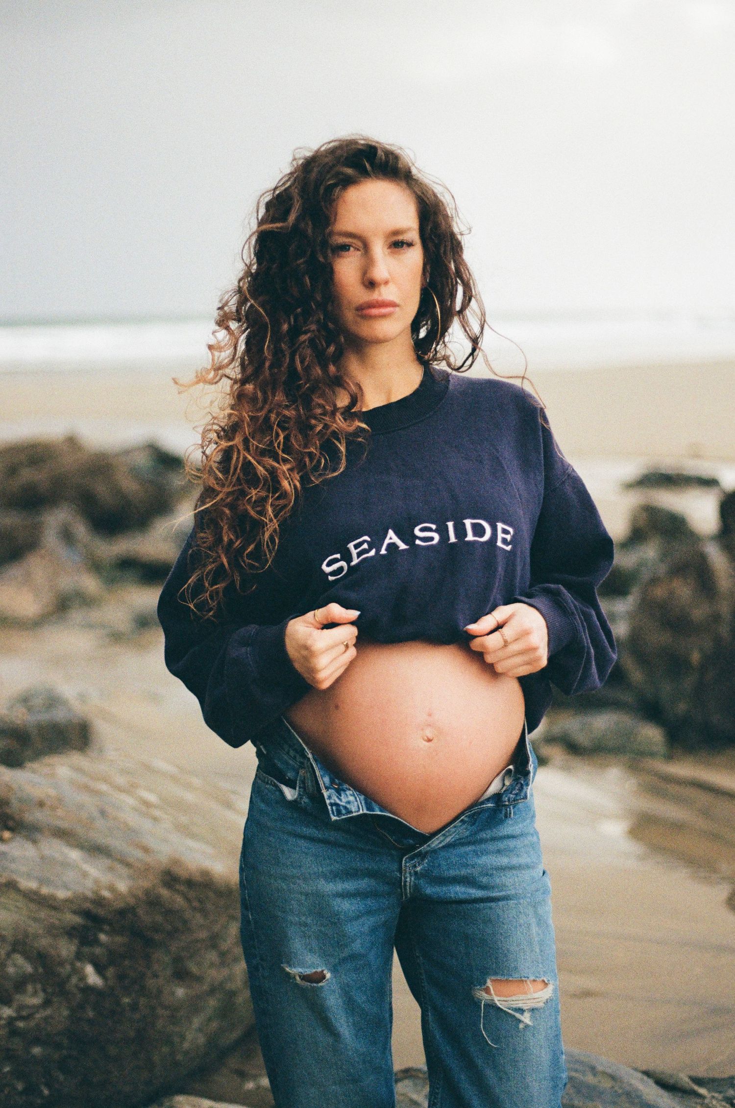 Maternity photos on a beach showing a pregnant belly under a navy Seaside sweatshirt with ripped jeans.