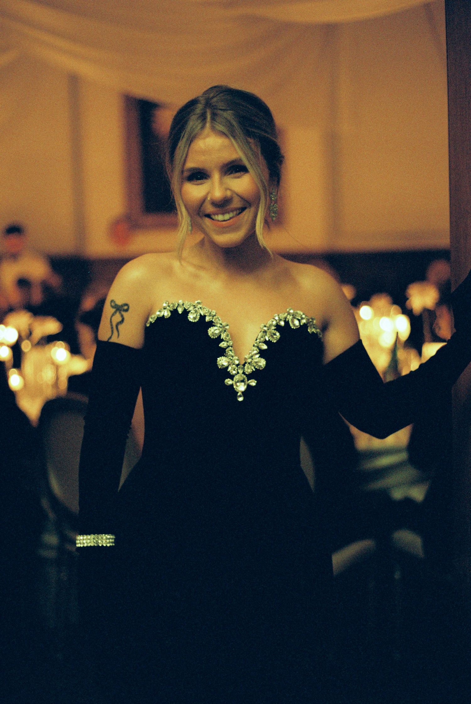 Person wearing a black off-shoulder dress poses in a dimly lit venue with warm ambient lighting in the background.