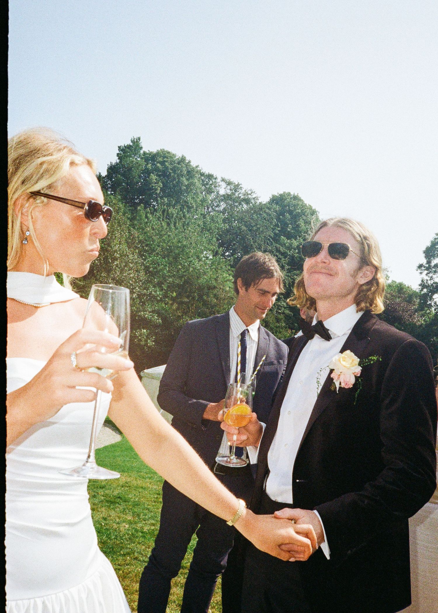 Wedding guests in formal attire toast with drinks outdoors on a sunny day.