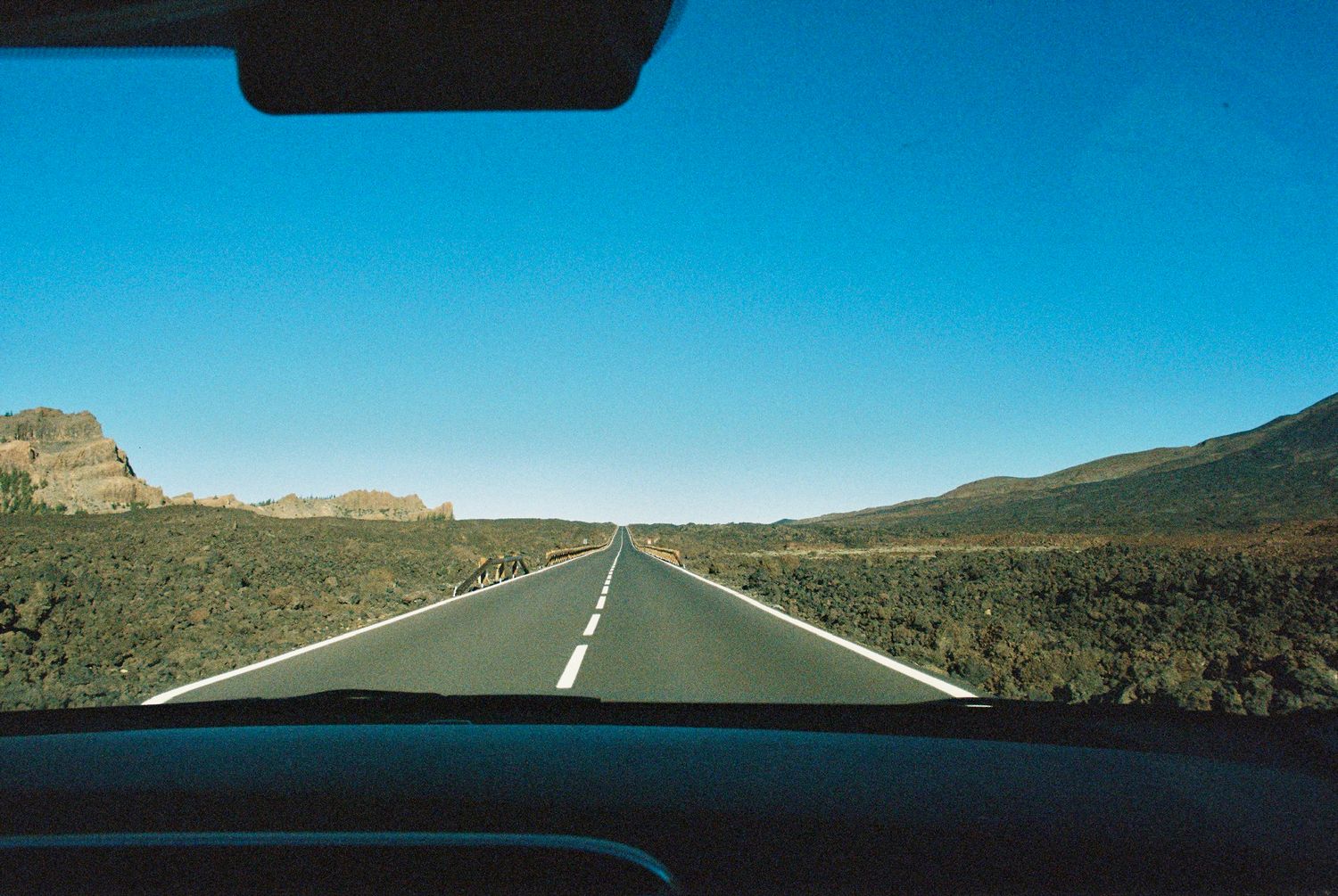 Empty highway stretches into distance through desert landscape under bright blue sky.