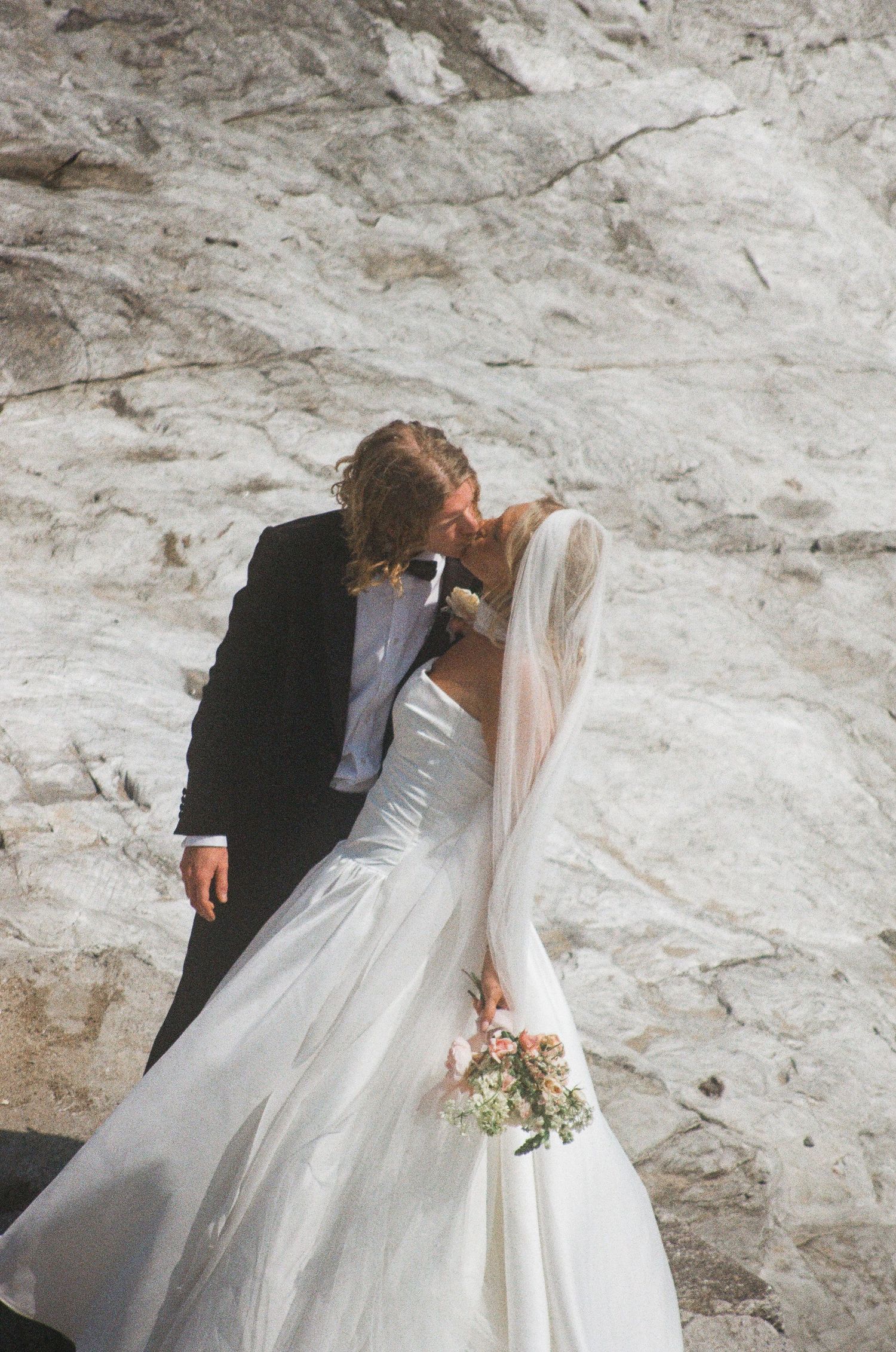 A romantic wedding moment captured against a textured white stone wall background.