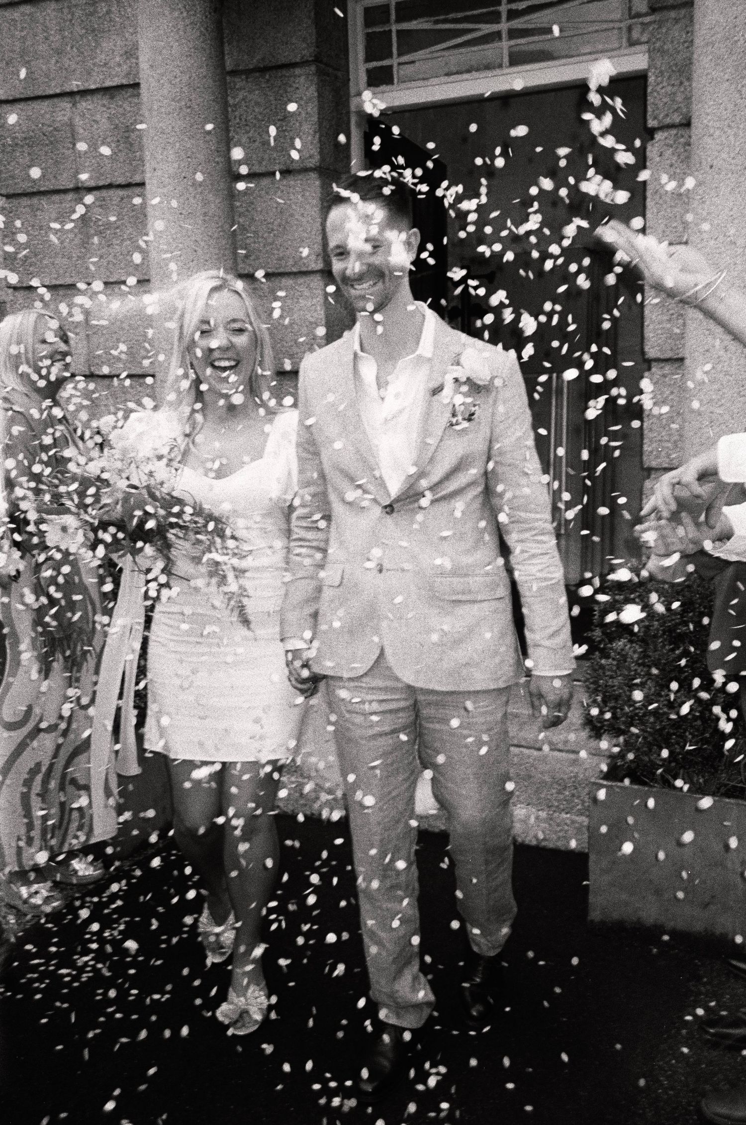 Black and white image of wedding party walking through a shower of confetti.