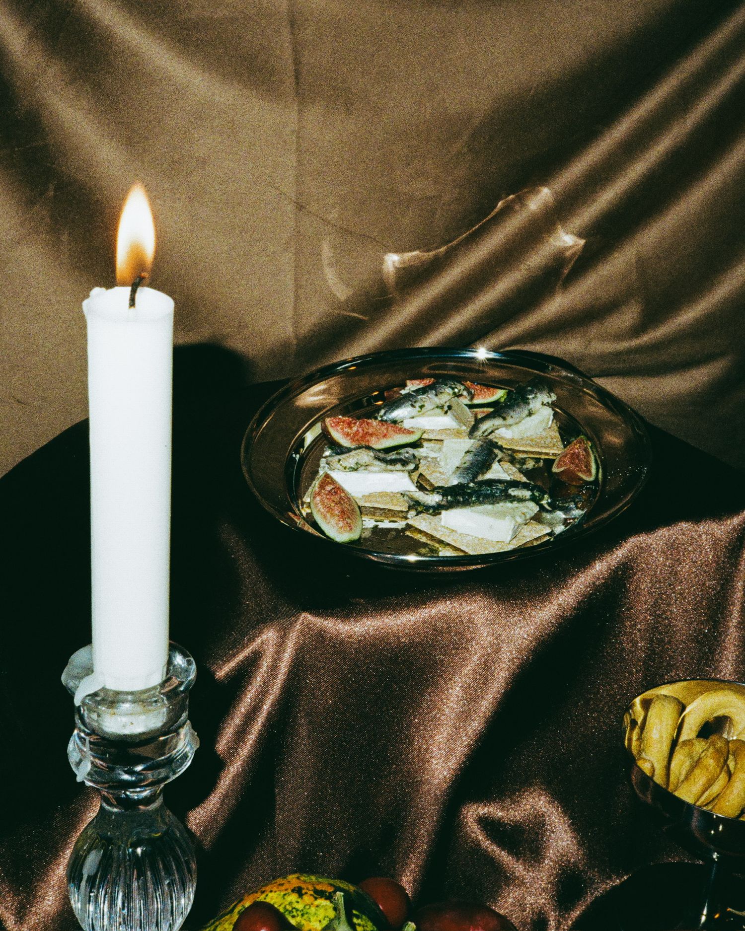 Lit white candles and decorative items arranged on a brown satin tablecloth.