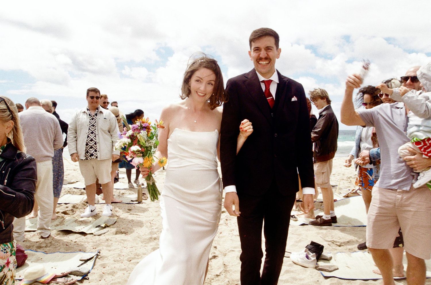 Beach wedding ceremony with couple and guests gathered on sandy shore with ocean waves in background.