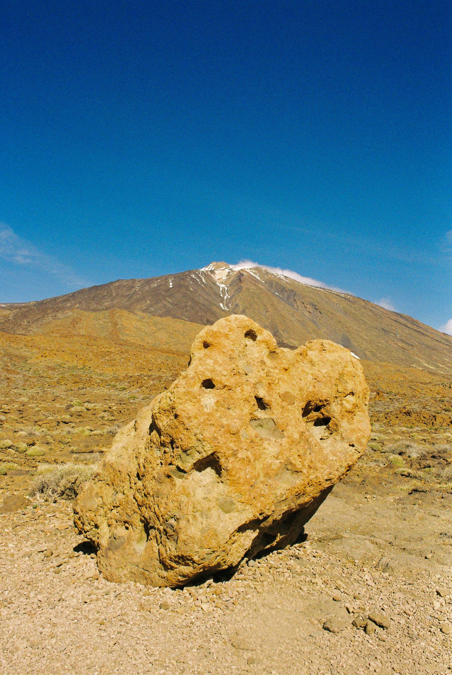 Large weathered rock formation with holes sits in a desert landscape with mountains and blue sky in the background.