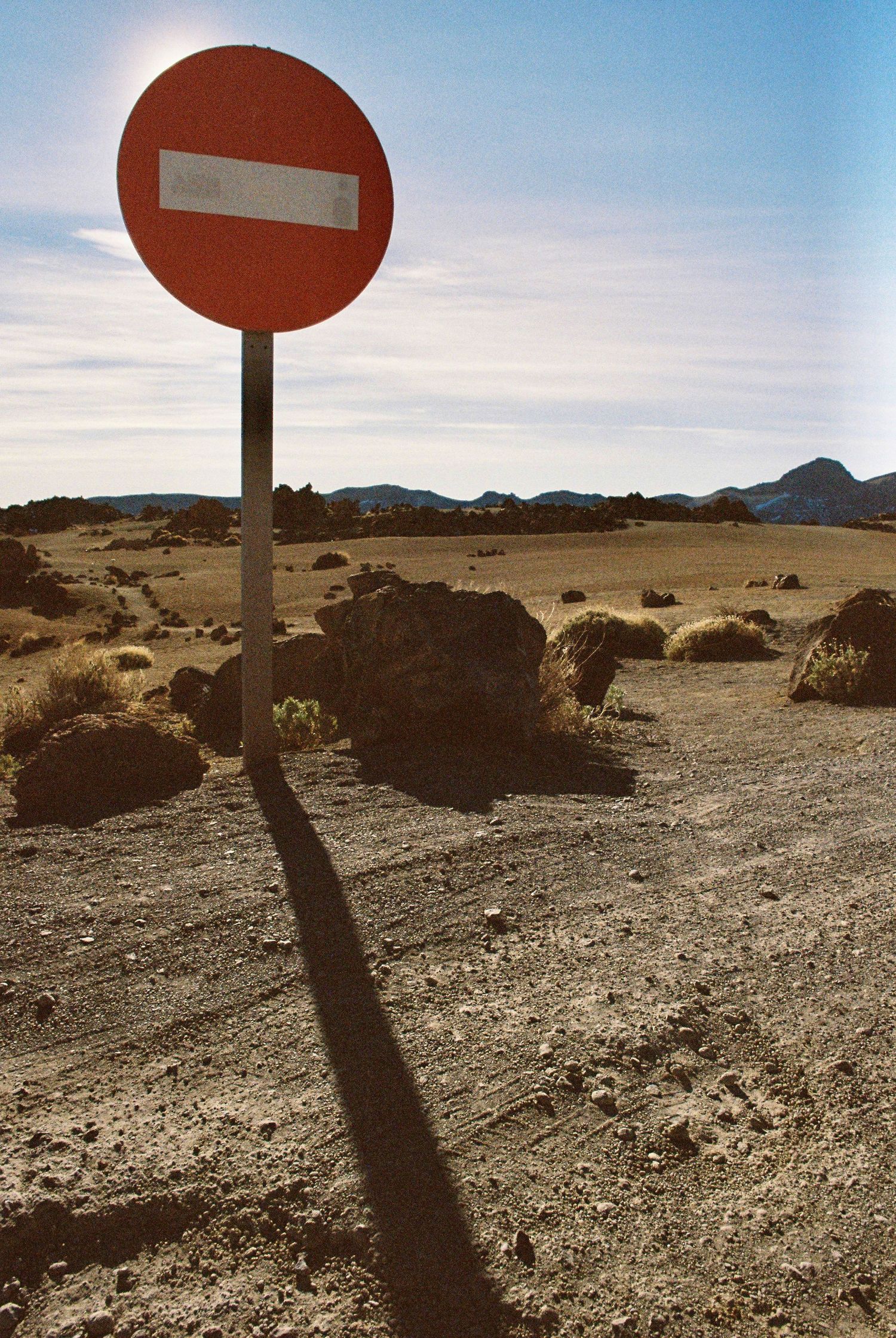 Red circular no entry traffic sign casting long shadow on rocky desert terrain.