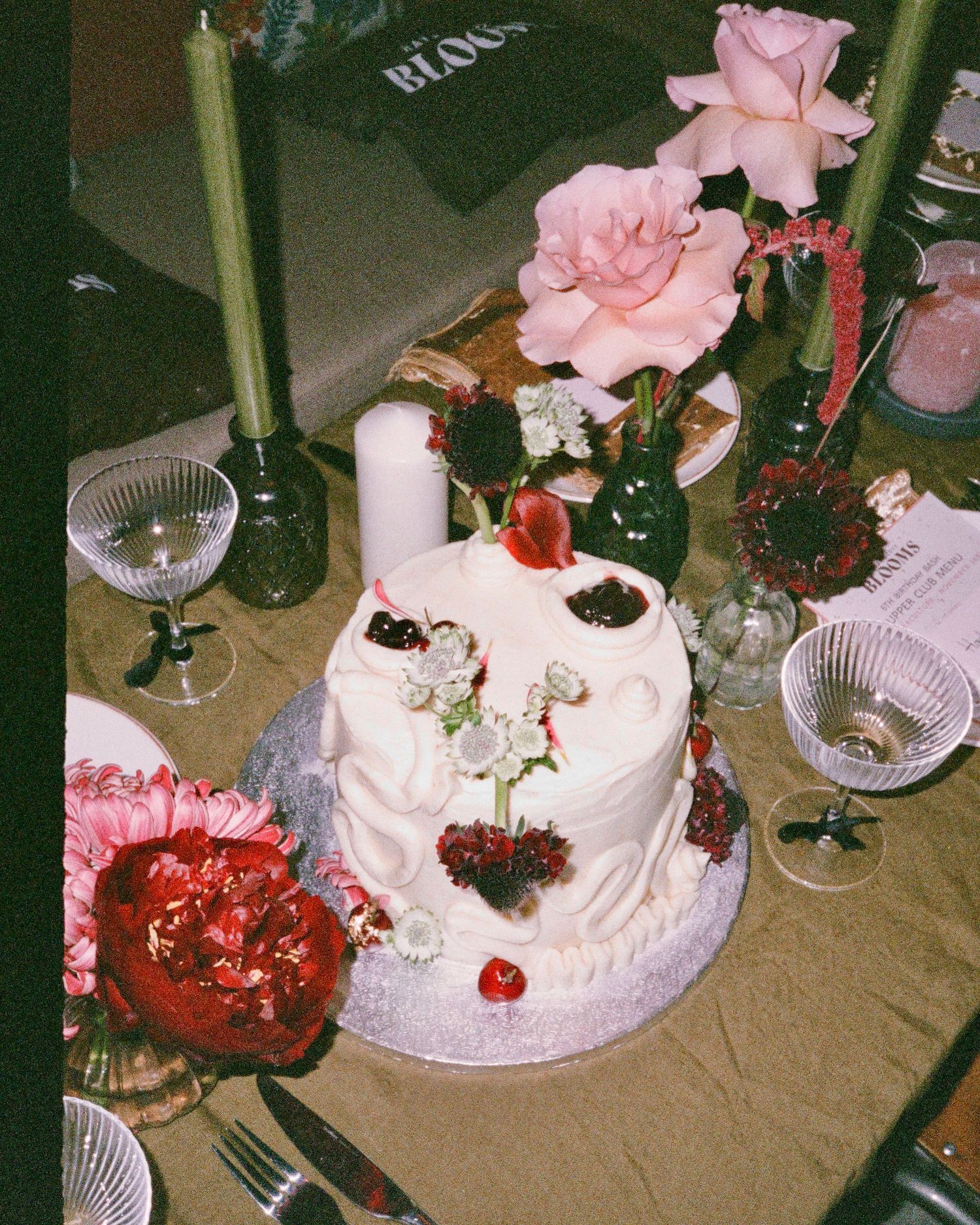 White wedding cake decorated with pink roses and red berries on a table with glassware.