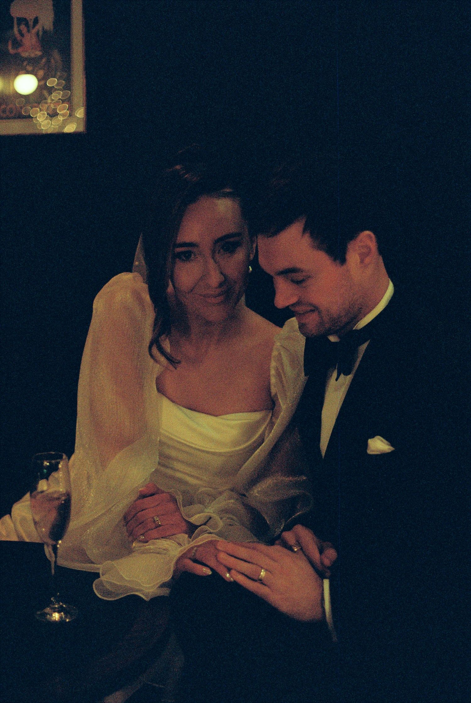 A couple in formal wedding attire share an intimate moment while looking at a ring in dim lighting.