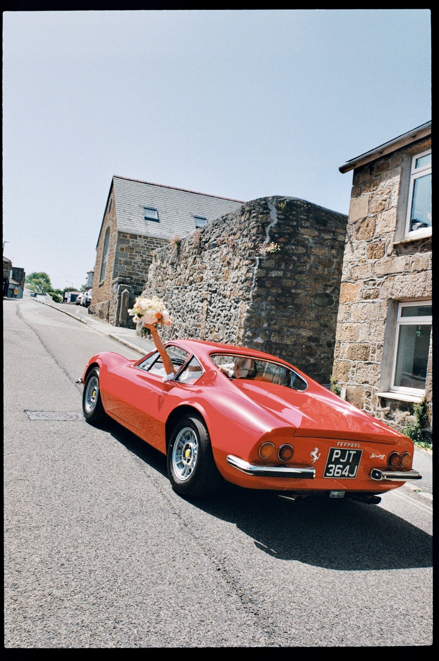 A classic red Ferrari Dino with a bridal bouquet on the roof parked on a sunny street beside stone buildings.