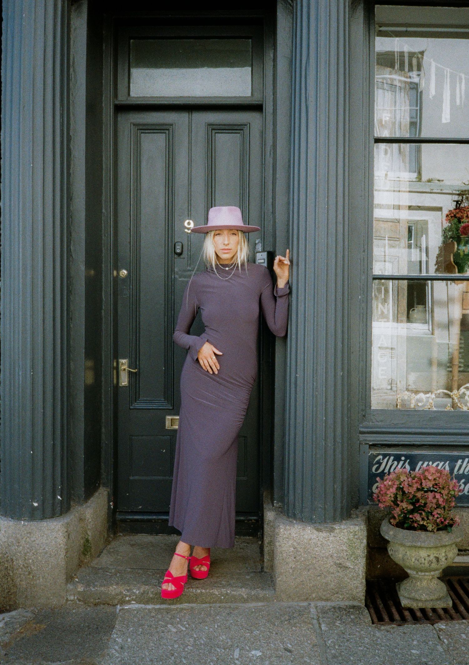 Playful fashion poses in purple dress and pink hat by a dark doorway.