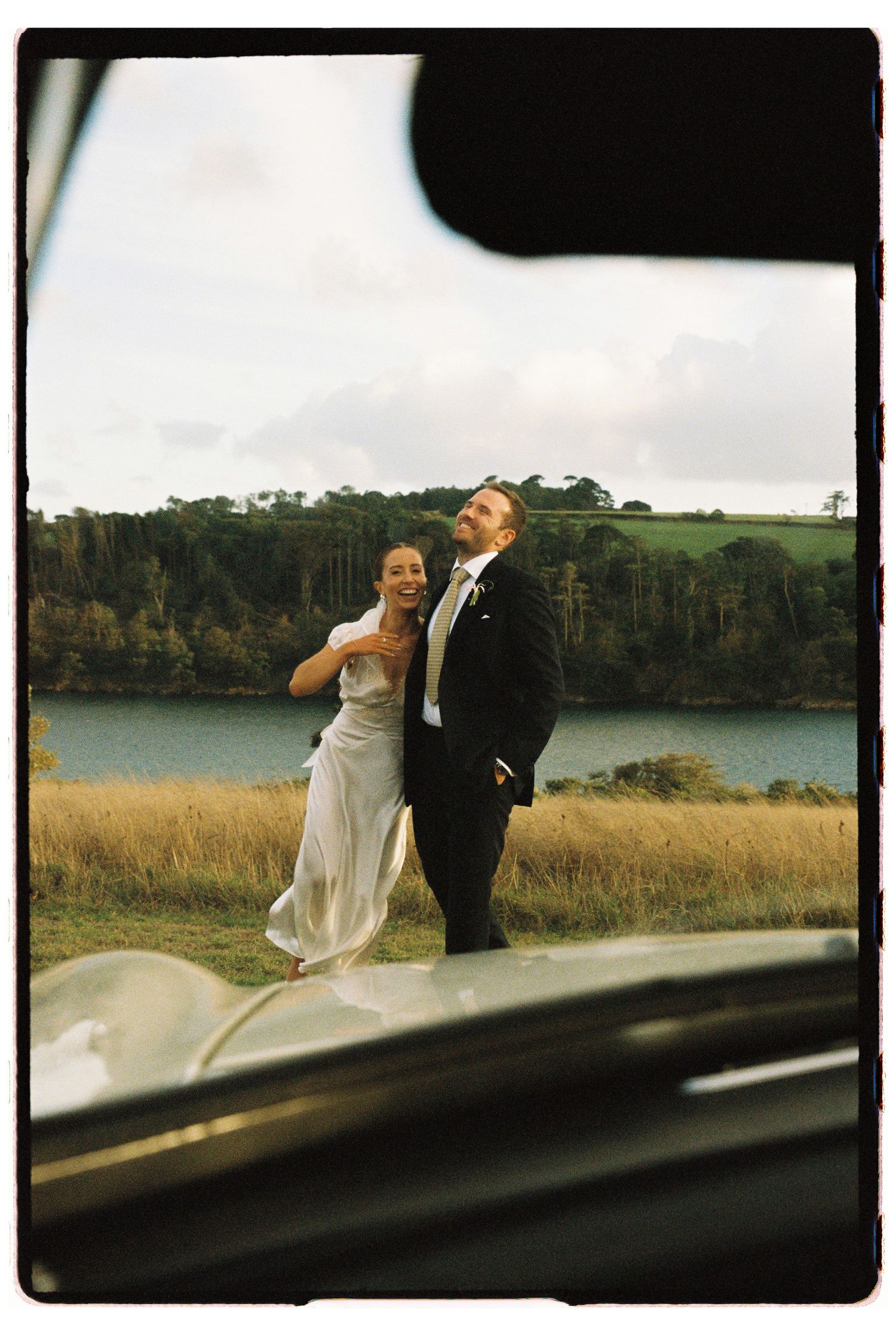 A bride and groom laugh joyfully beside a lake and golden fields, shot through a car window on film.