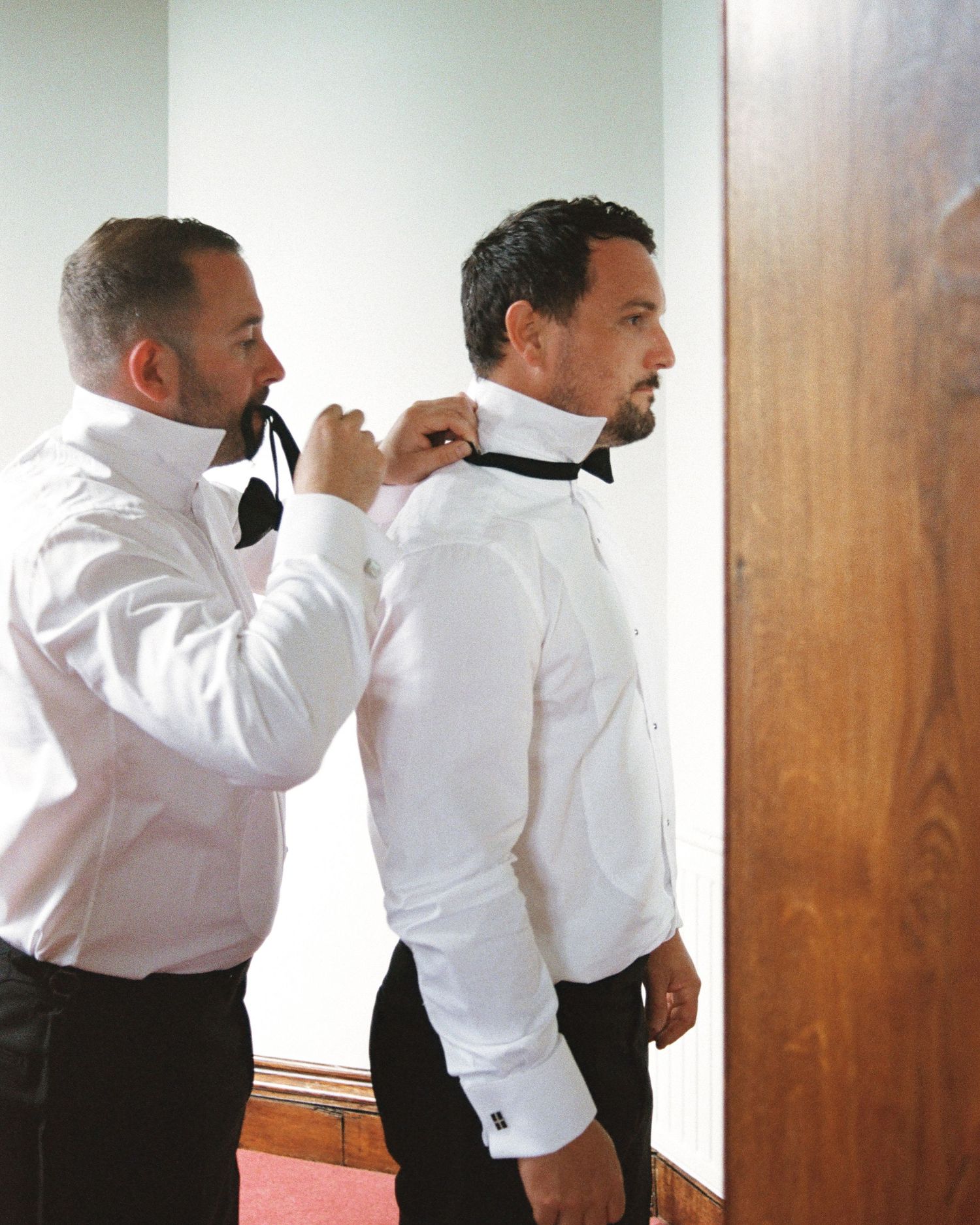 Groomsmen helping each other get dressed in white shirts before wedding ceremony.