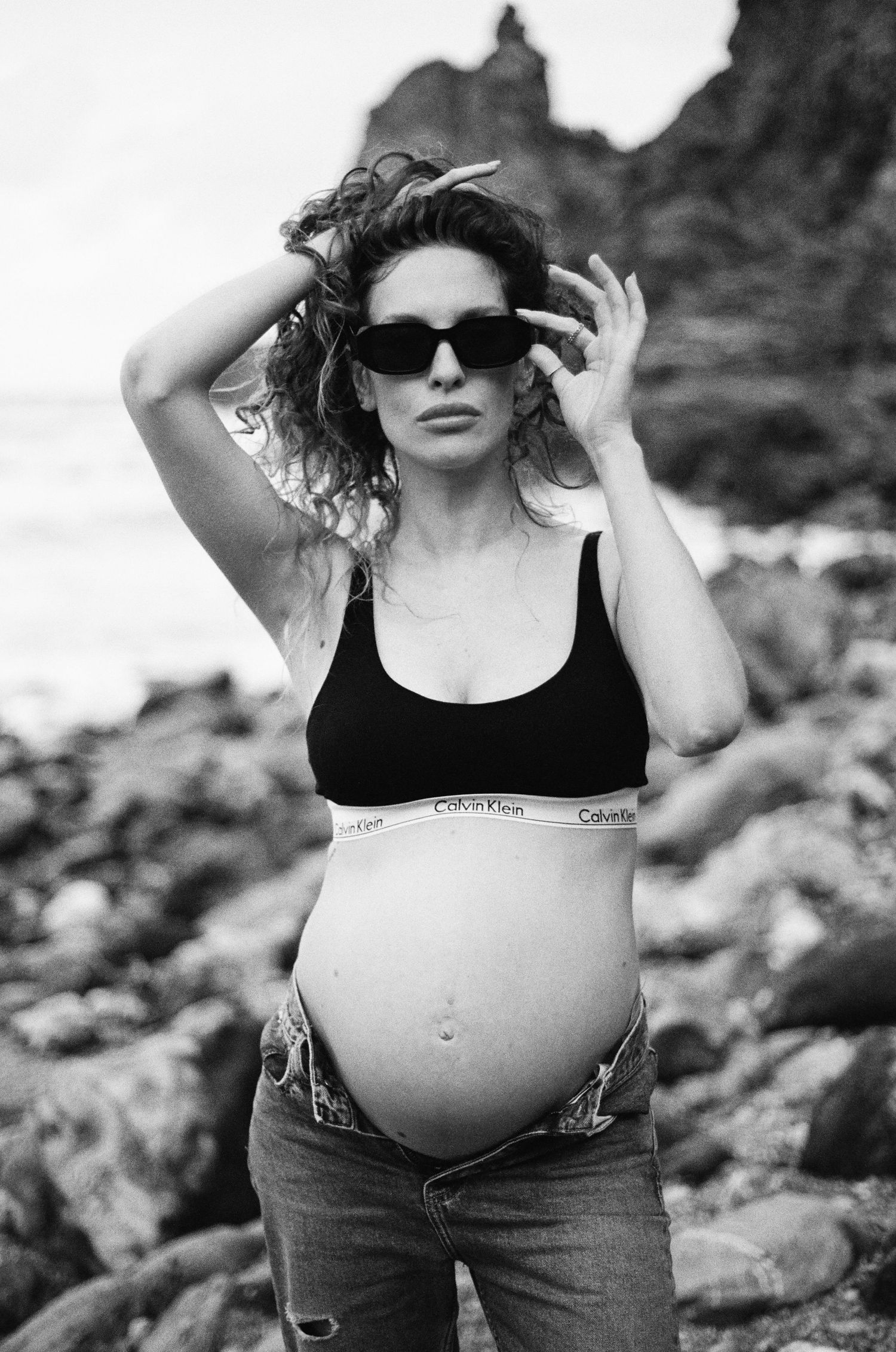 Black and white maternity photos on rocky coastline showing casual style and sunglasses.