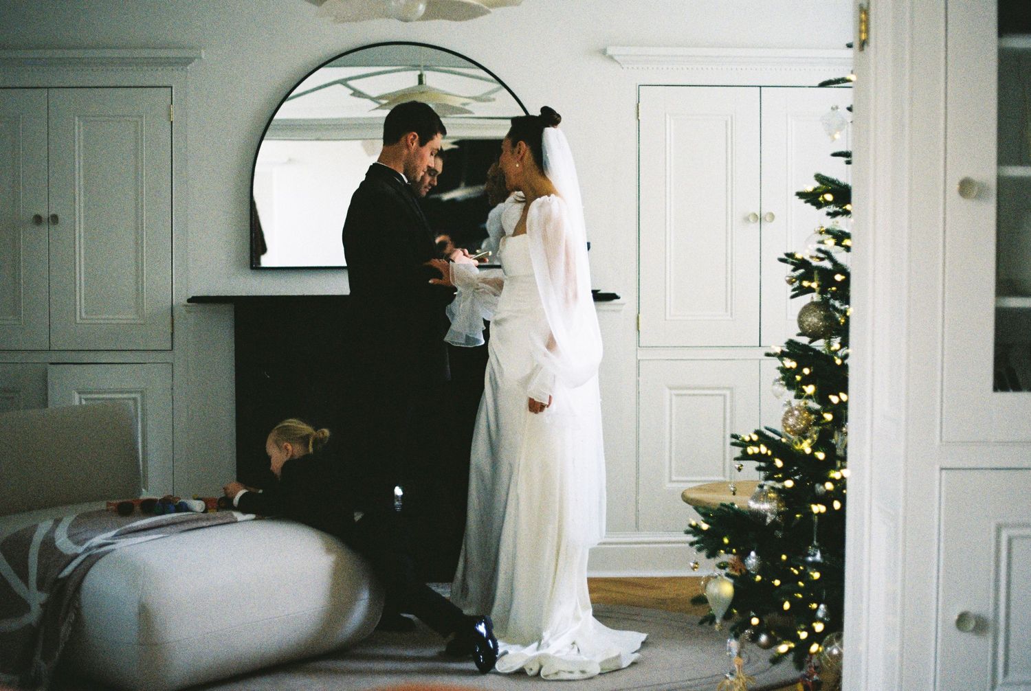 A couple shares an intimate moment in a bright white room decorated for Christmas with a tree in the corner.