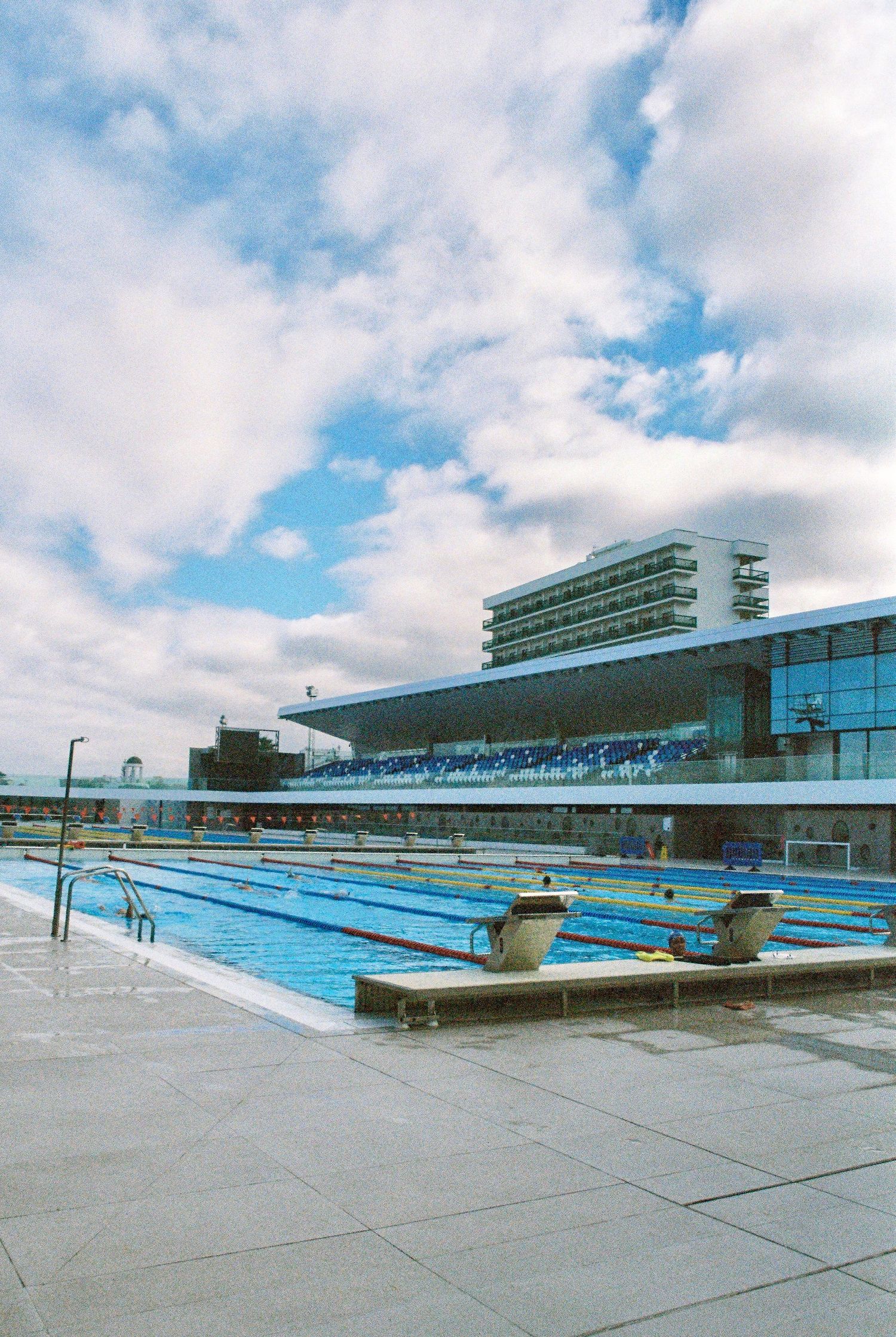 Modern outdoor swimming pool complex with multiple lanes and spectator stands under cloudy sky.