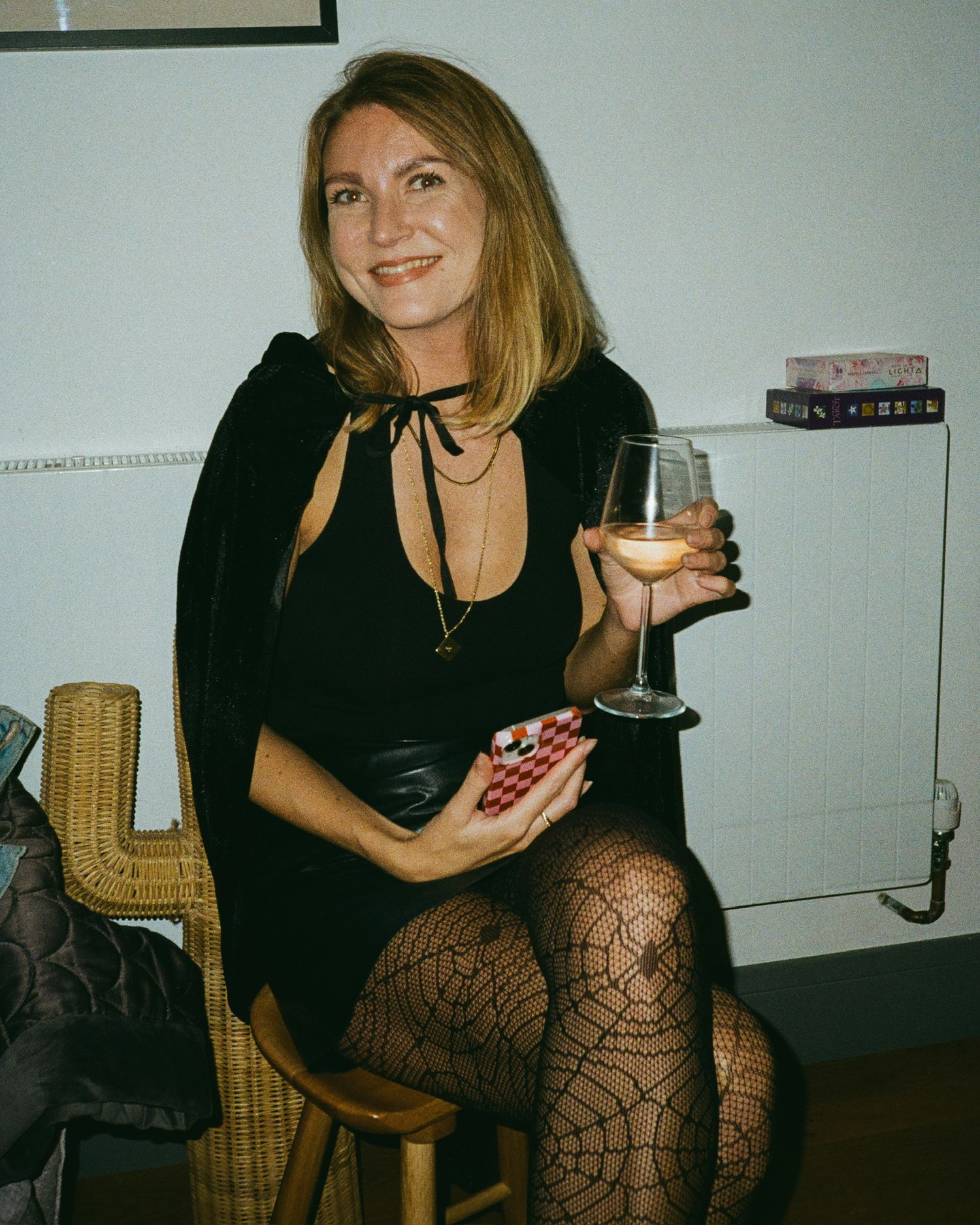 A person in black attire sits in a wicker chair holding a drink glass in a dimly lit room.