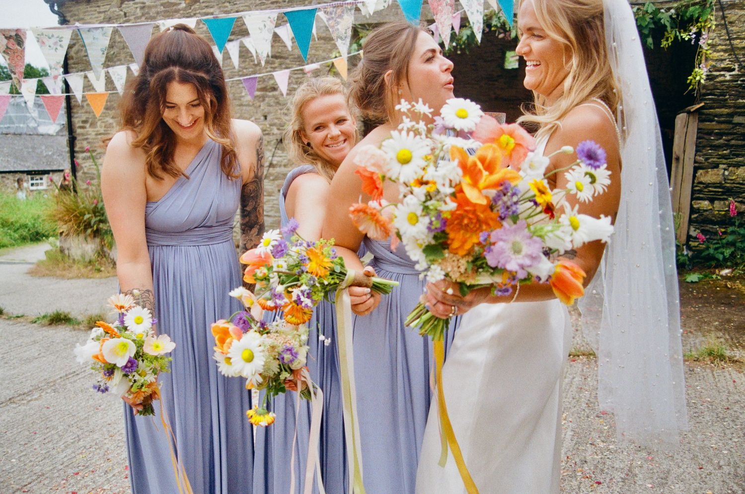 Bridesmaids in lavender dresses hold vibrant orange and pink bouquets outdoors.