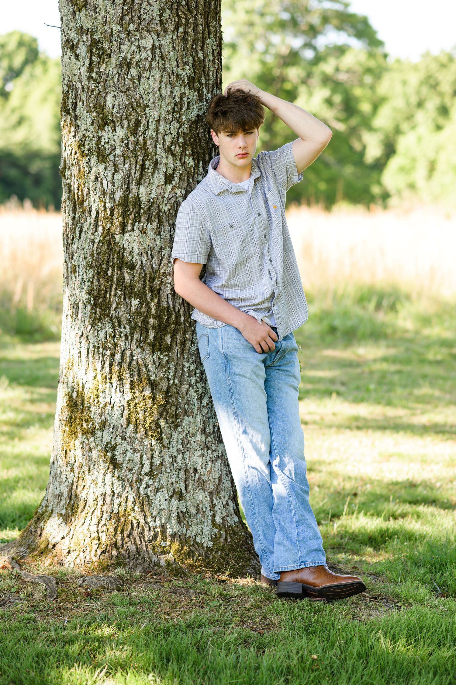 Young man in jeans and gray shirt leaning against a large tree trunk in a sunny green park.
