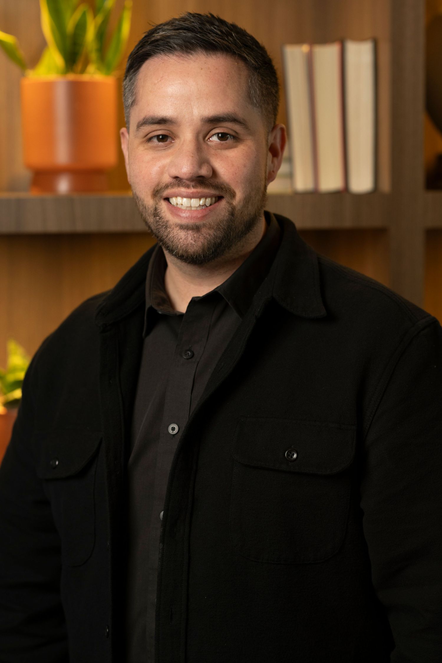 Corporate headshot of person wearing all black attire against blurred office background with plants.