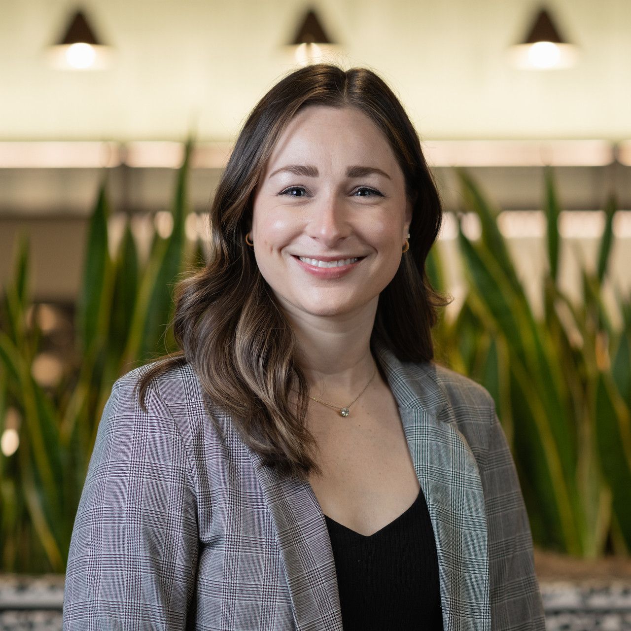 Professional headshot of person wearing gray blazer and black top in office setting with plants in background.