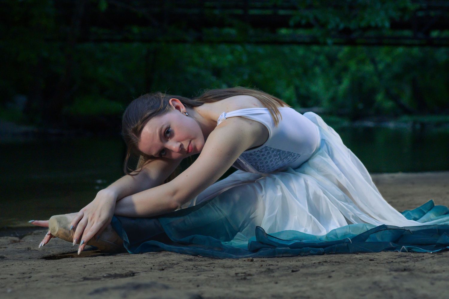 A young woman in a white and blue dress lies gracefully on sandy ground near a forest river.