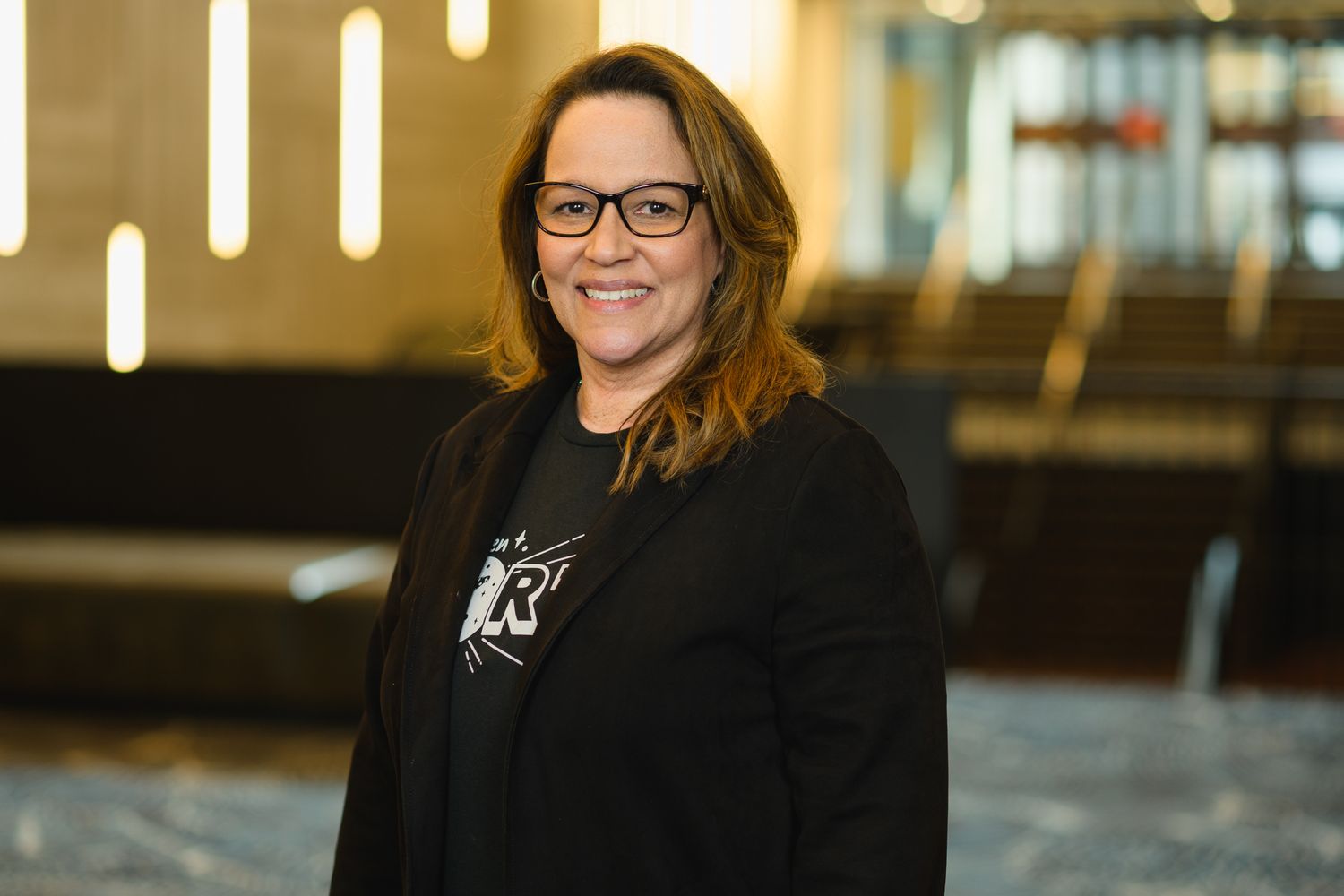 Professional headshot of someone wearing glasses and a dark shirt with company branding in an office setting.